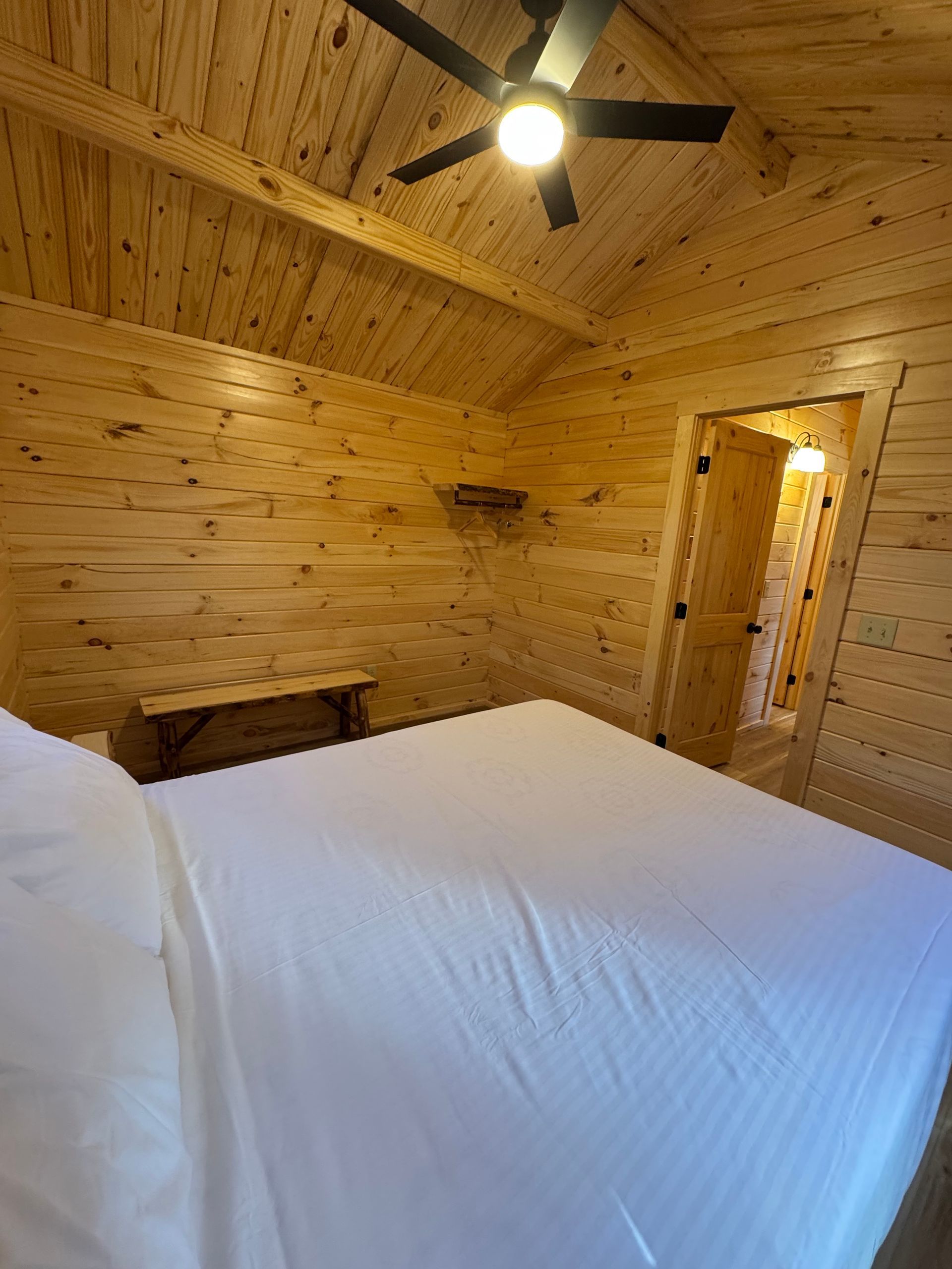 A bedroom featuring light wood paneled walls, a ceiling fan, a made bed with white linens, and an open doorway to a bathroom.