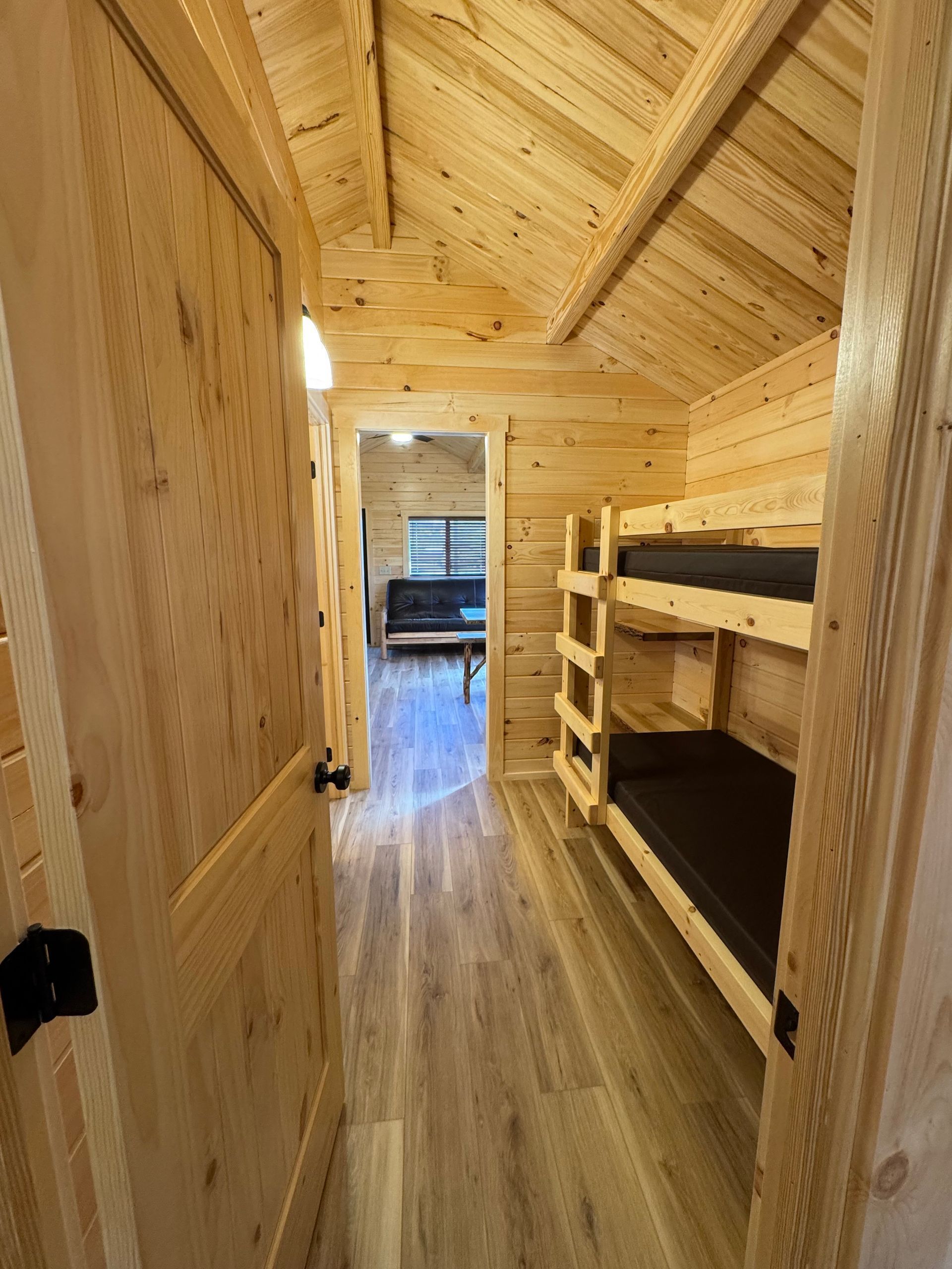 A view through a wooden doorway into a small, pine-walled cabin room with bunk beds and a hallway leading to a sofa.