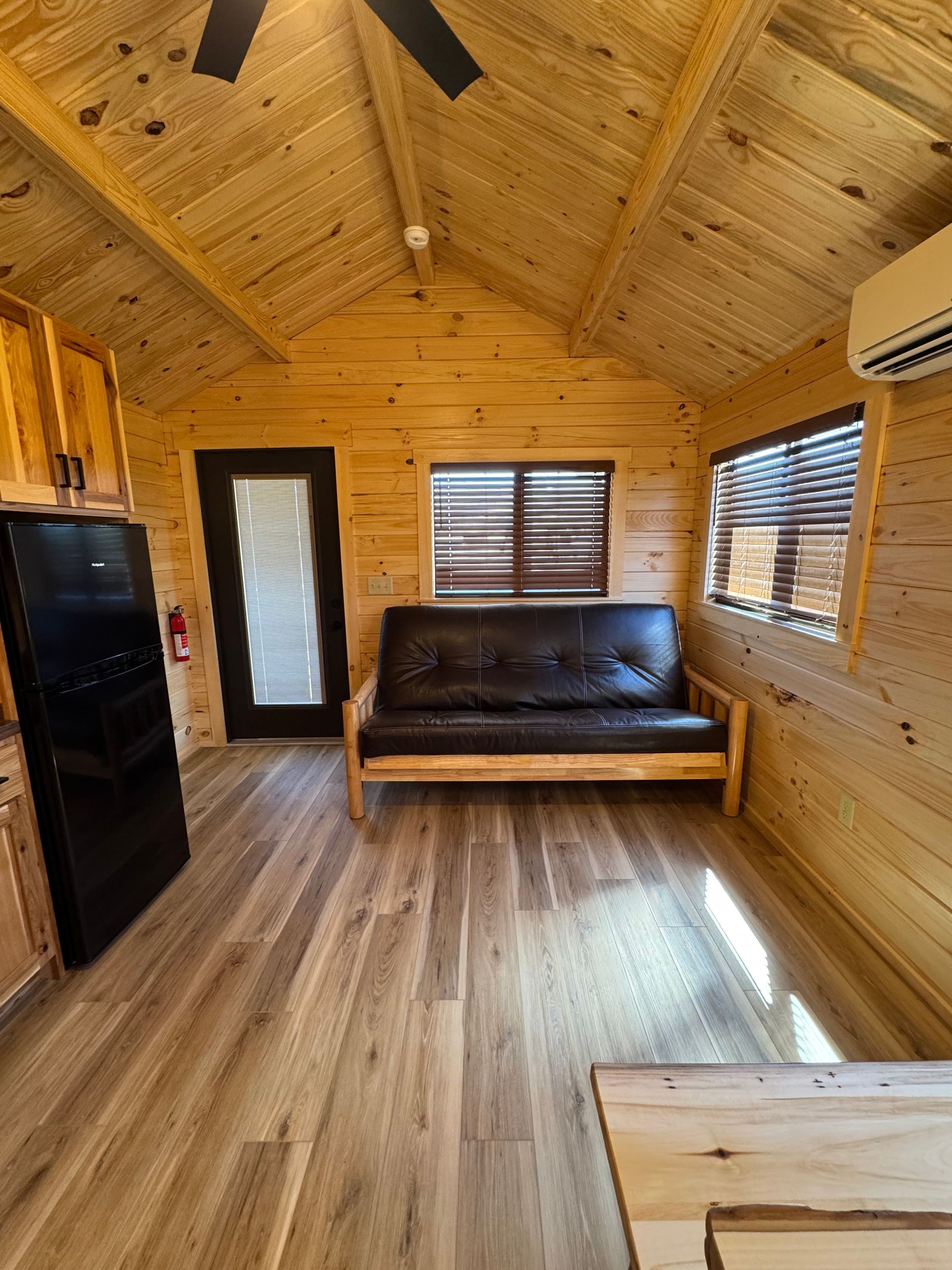 A small, wood-paneled cabin interior featuring a dark sofa, a black refrigerator, and a window with blinds.