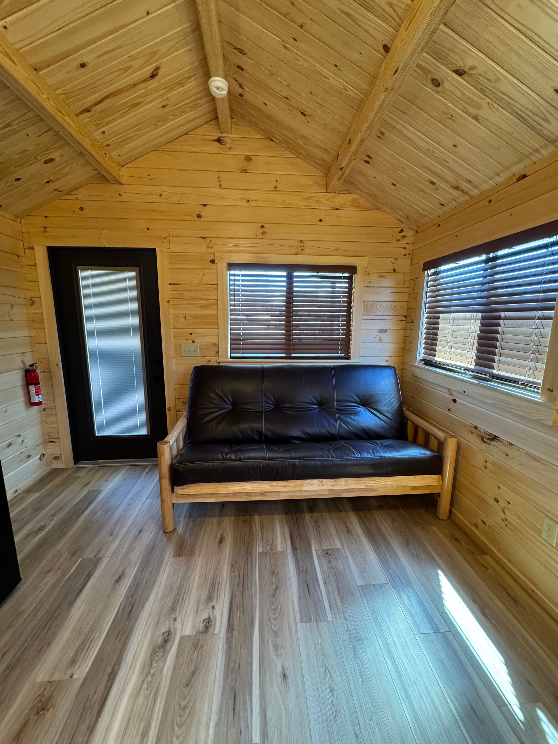 A small cabin interior with light wood walls, a dark futon, a black door, and two windows with closed wooden blinds.