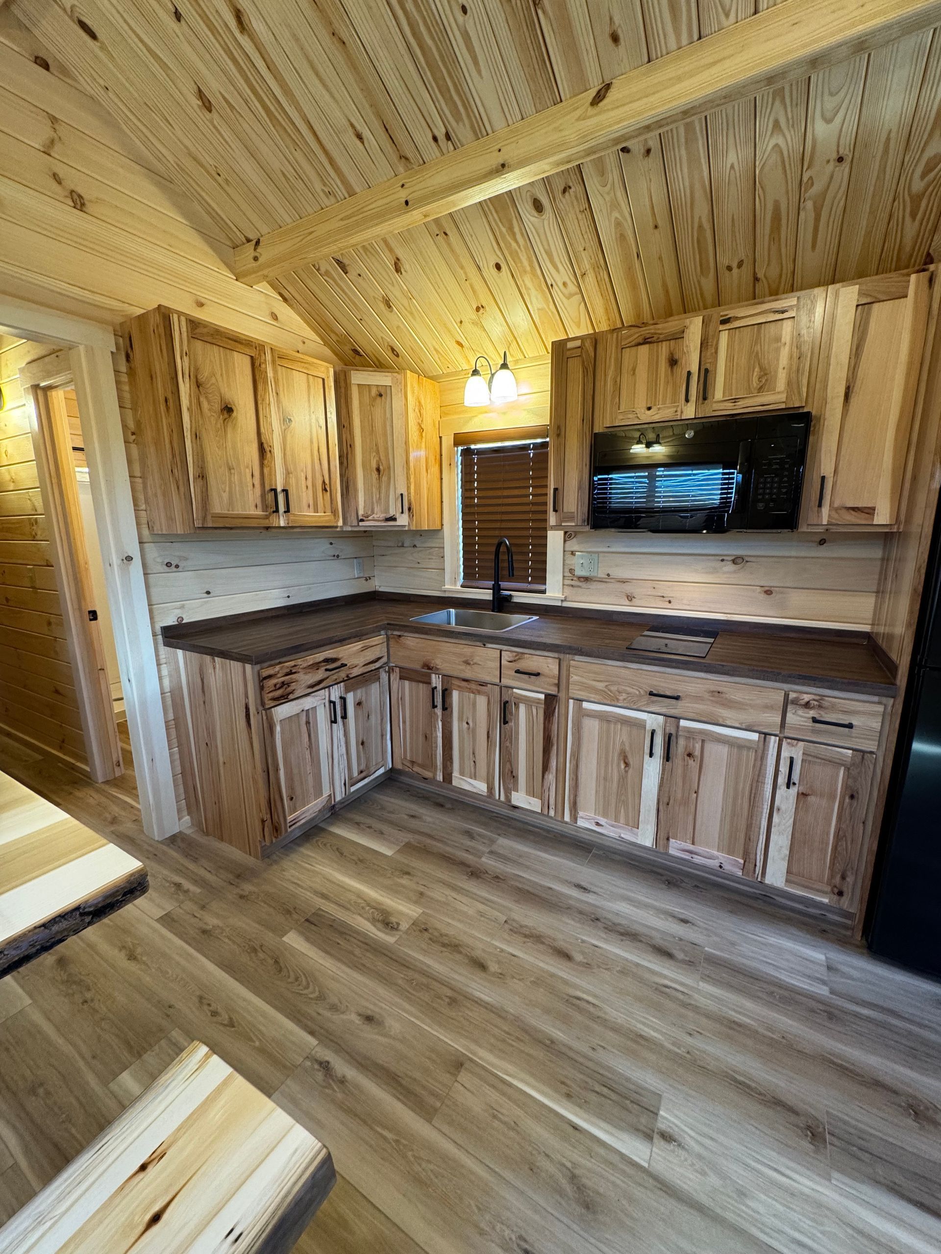 Rustic kitchen with light-wood knotty pine cabinetry, a dark countertop, and matching wood-paneled walls and ceiling.