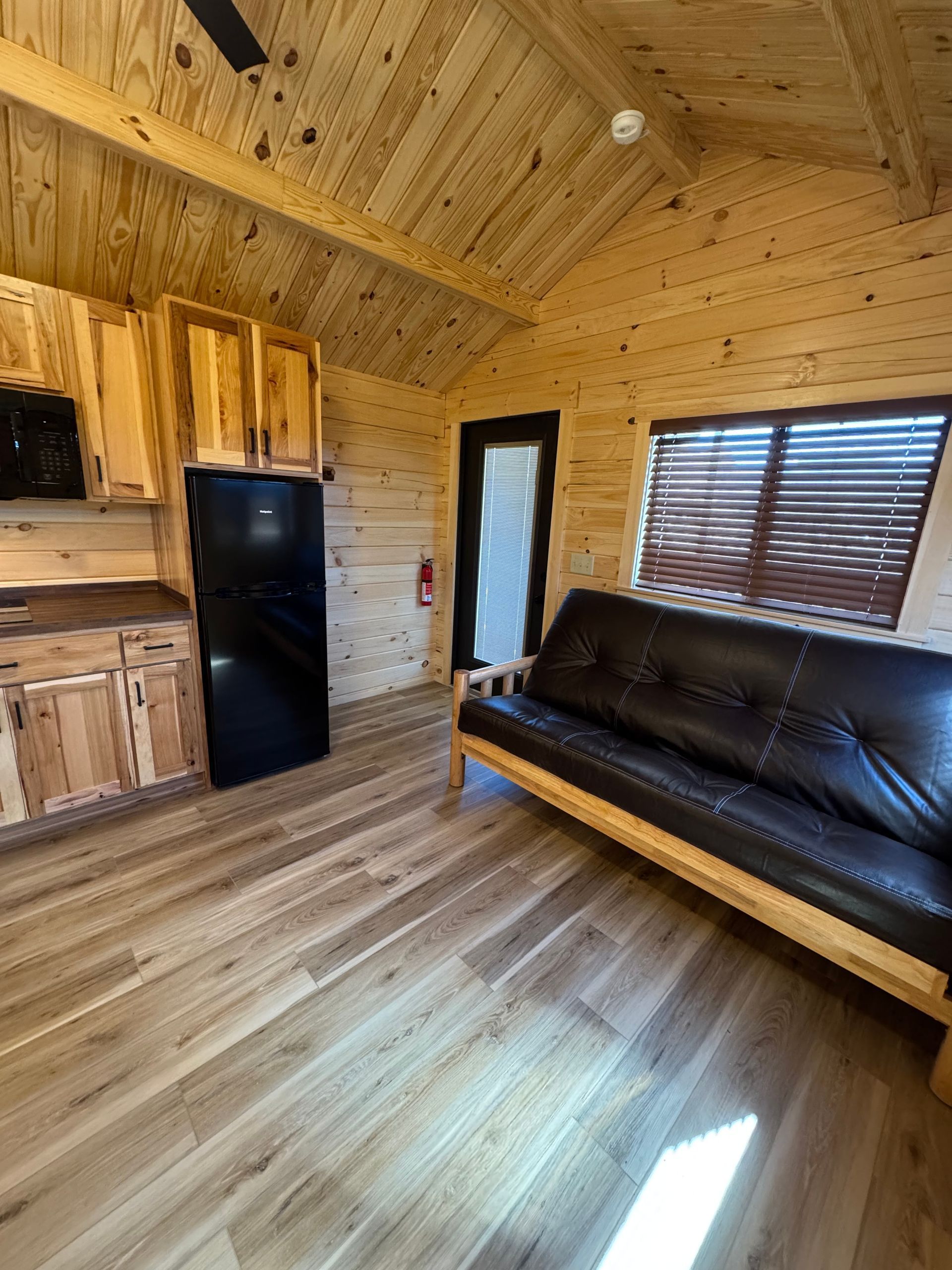 A small, wood-paneled cabin interior with a black futon, refrigerator, wooden cabinets, and a window with blinds.