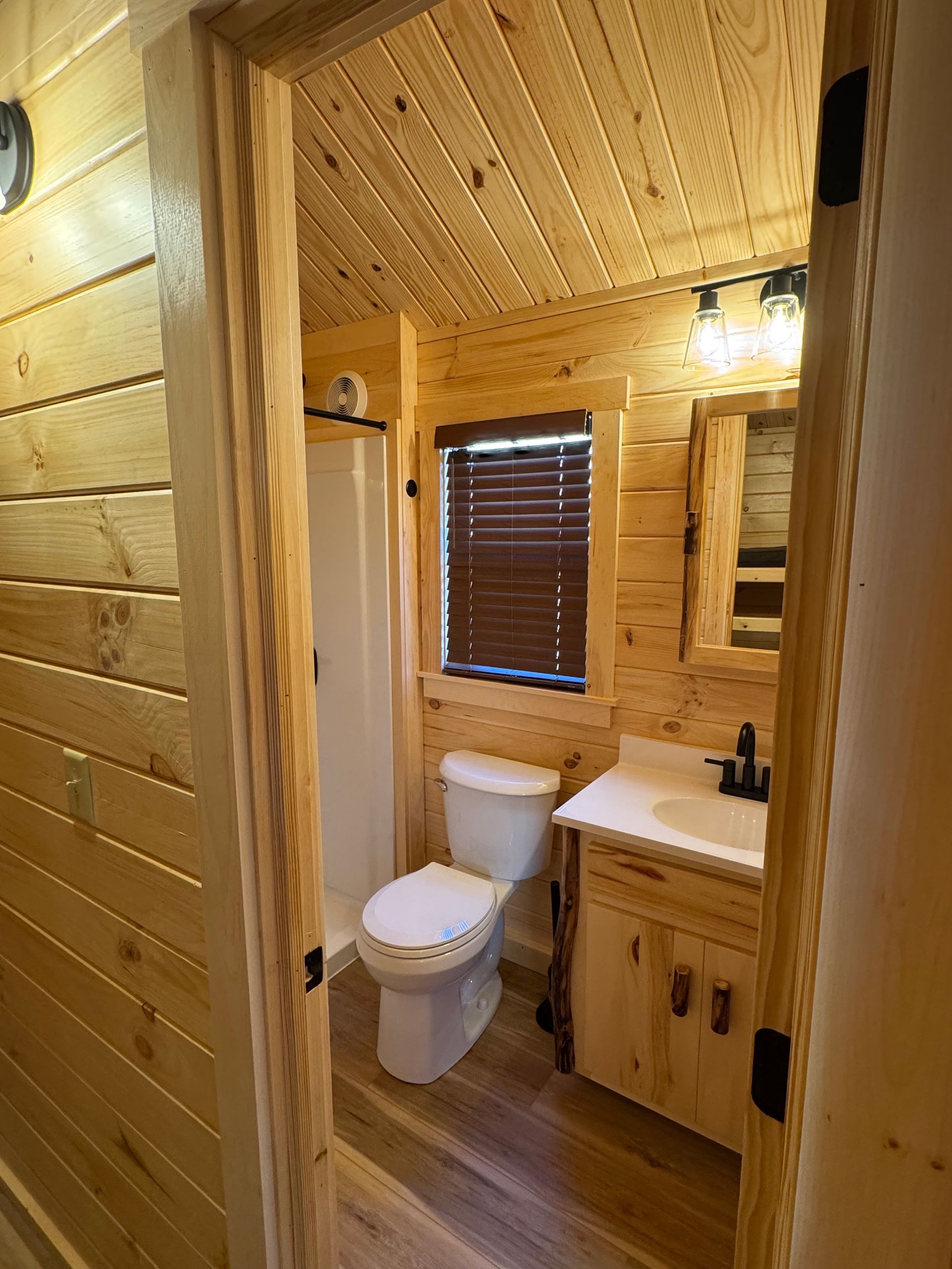 A bathroom with light wood paneling, a white toilet, a vanity with a sink, a mirror, and a shower stall.