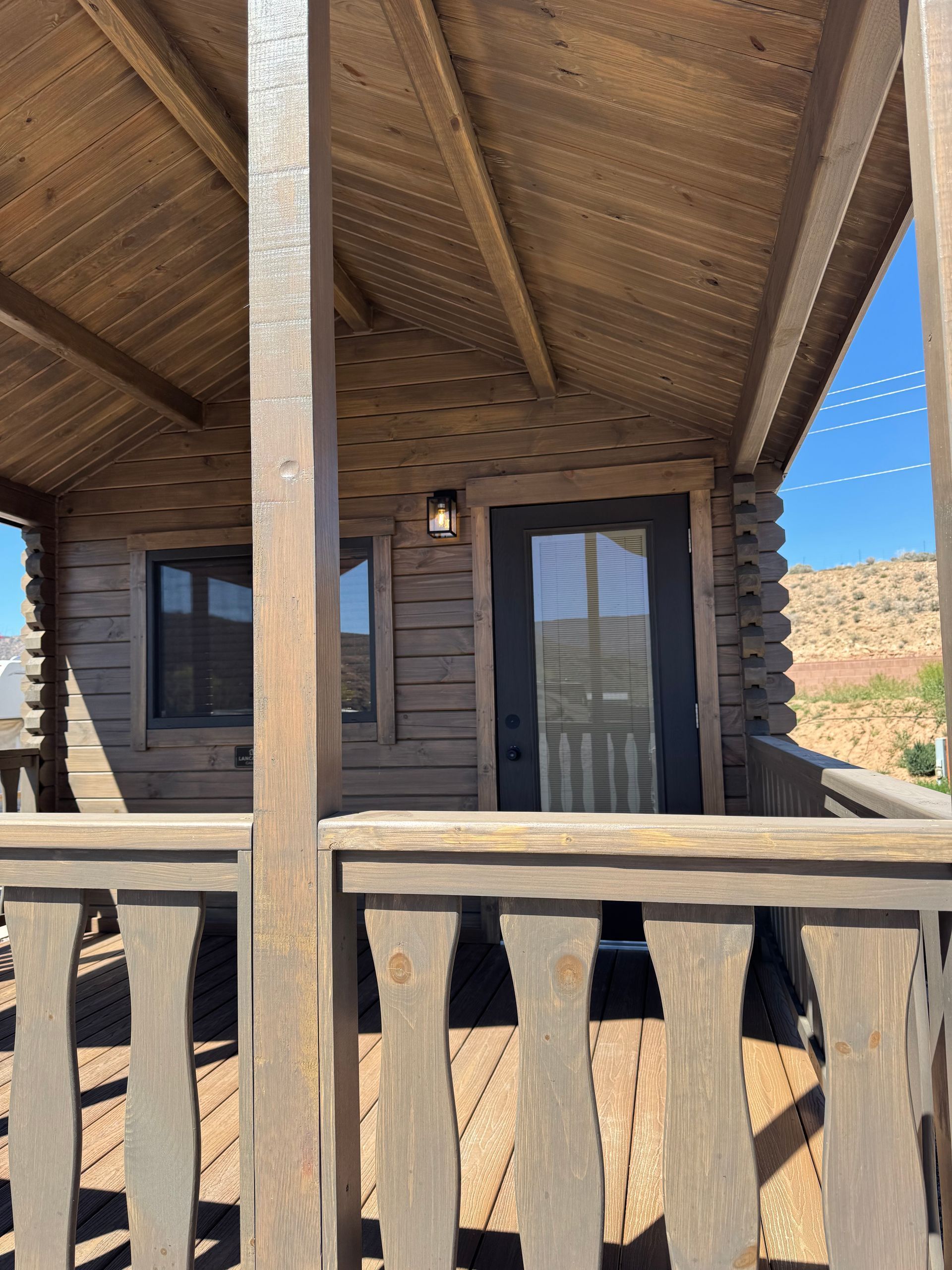 A wooden cabin porch with a window and a black door under a roof, overlooking a distant desert landscape.