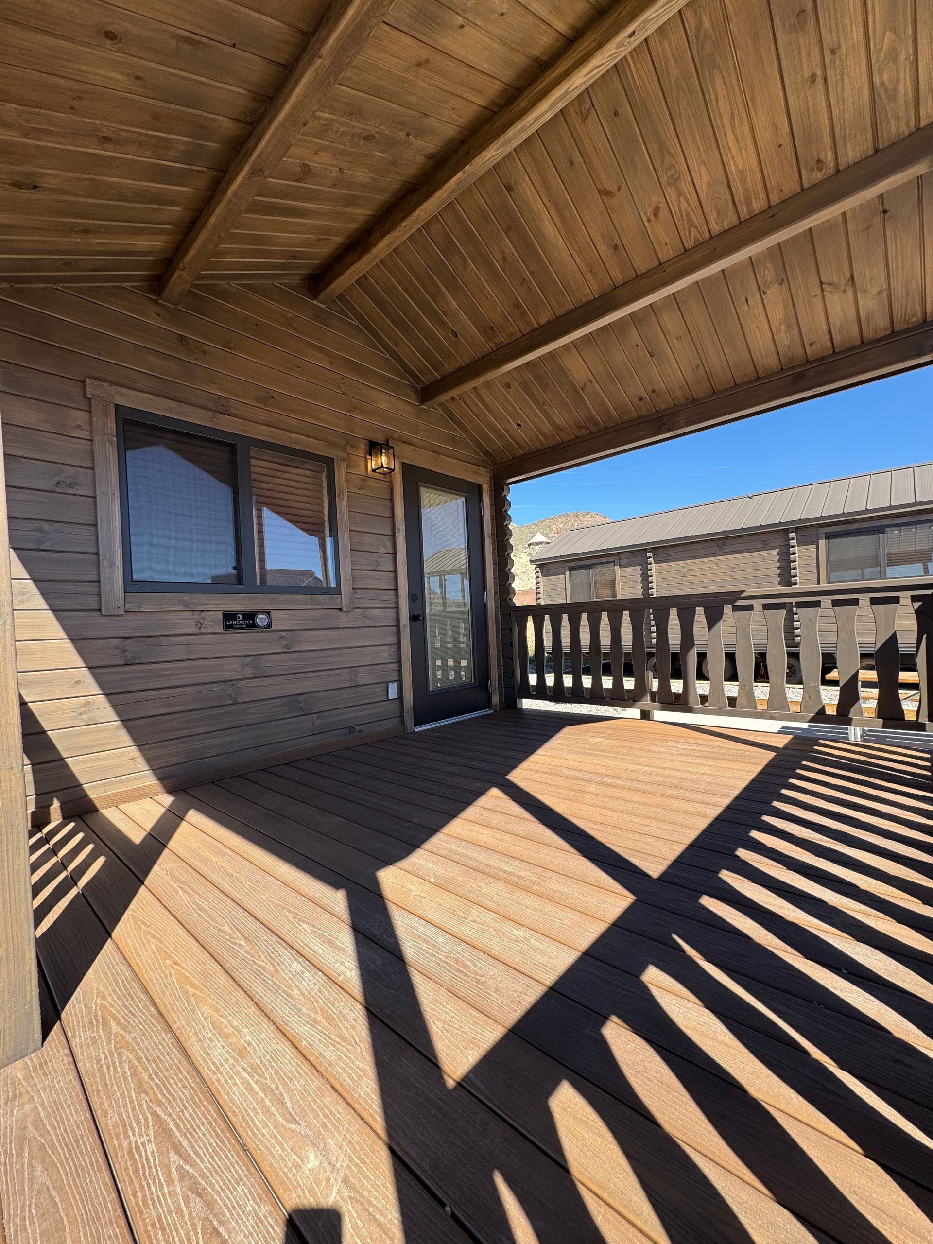 A wooden covered deck featuring a doorway, a window, and a wooden railing under a clear blue sky.