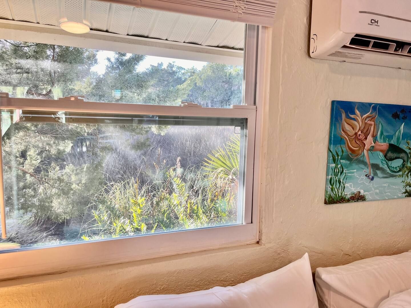 A bedroom with a large window and a painting of a mermaid on the wall.