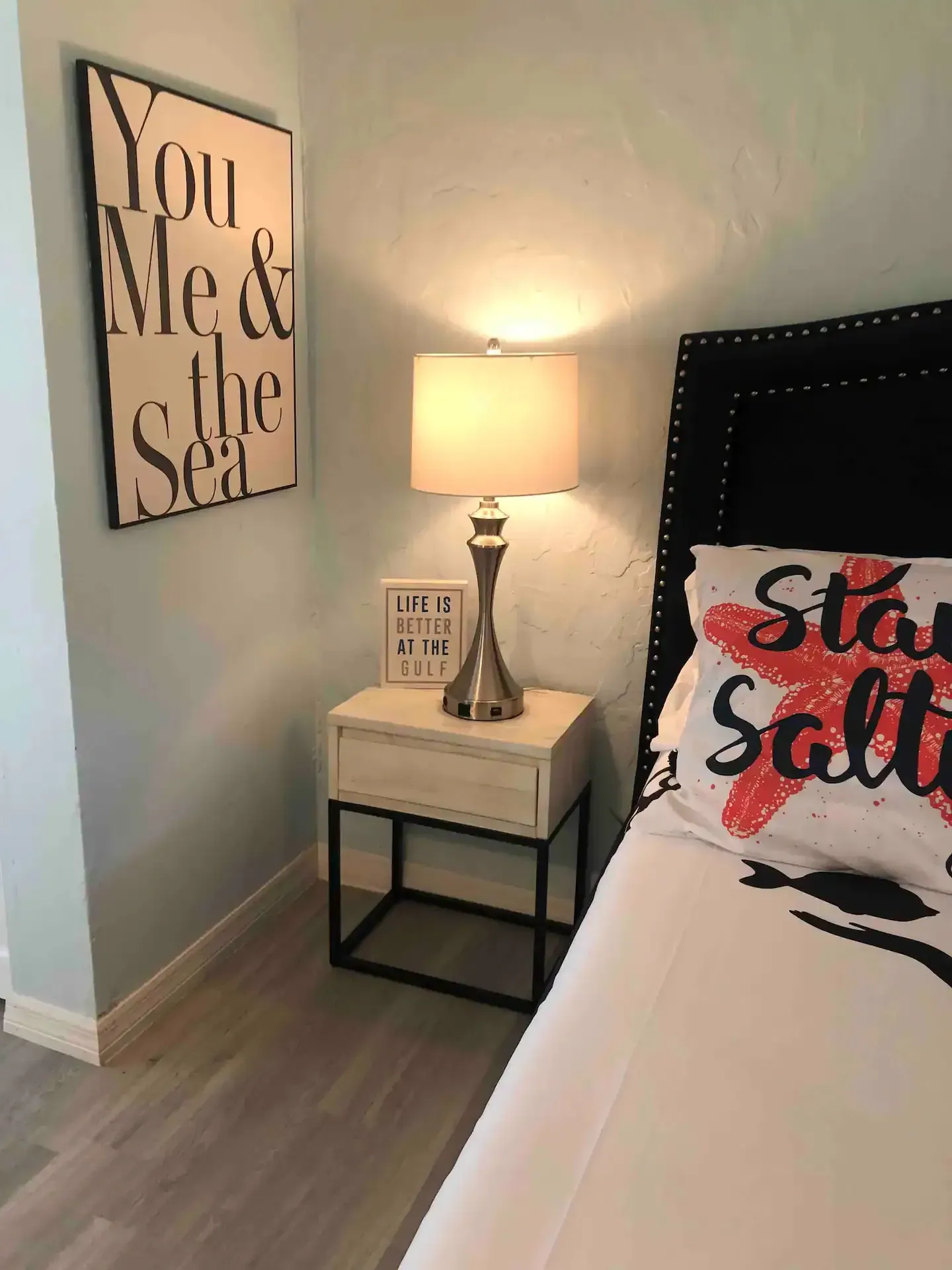 A bedroom with a bed , nightstand , lamp and a sign that says `` you me & the sea ''.