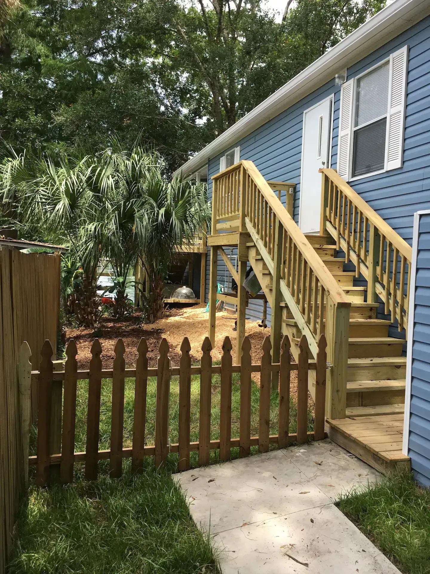A blue house with a wooden deck and stairs behind a picket fence.