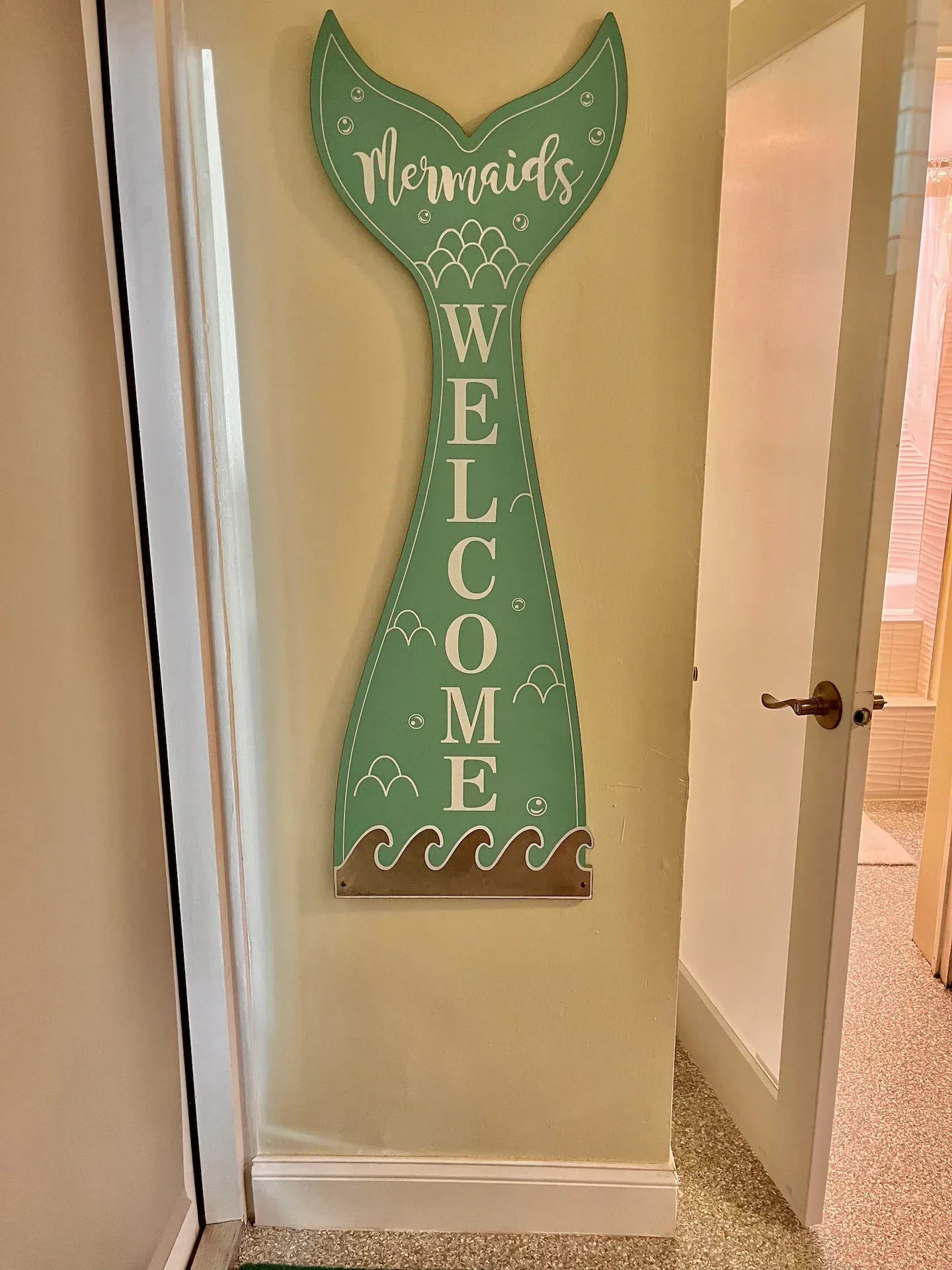 A green mermaid tail welcome sign is hanging on a wall.