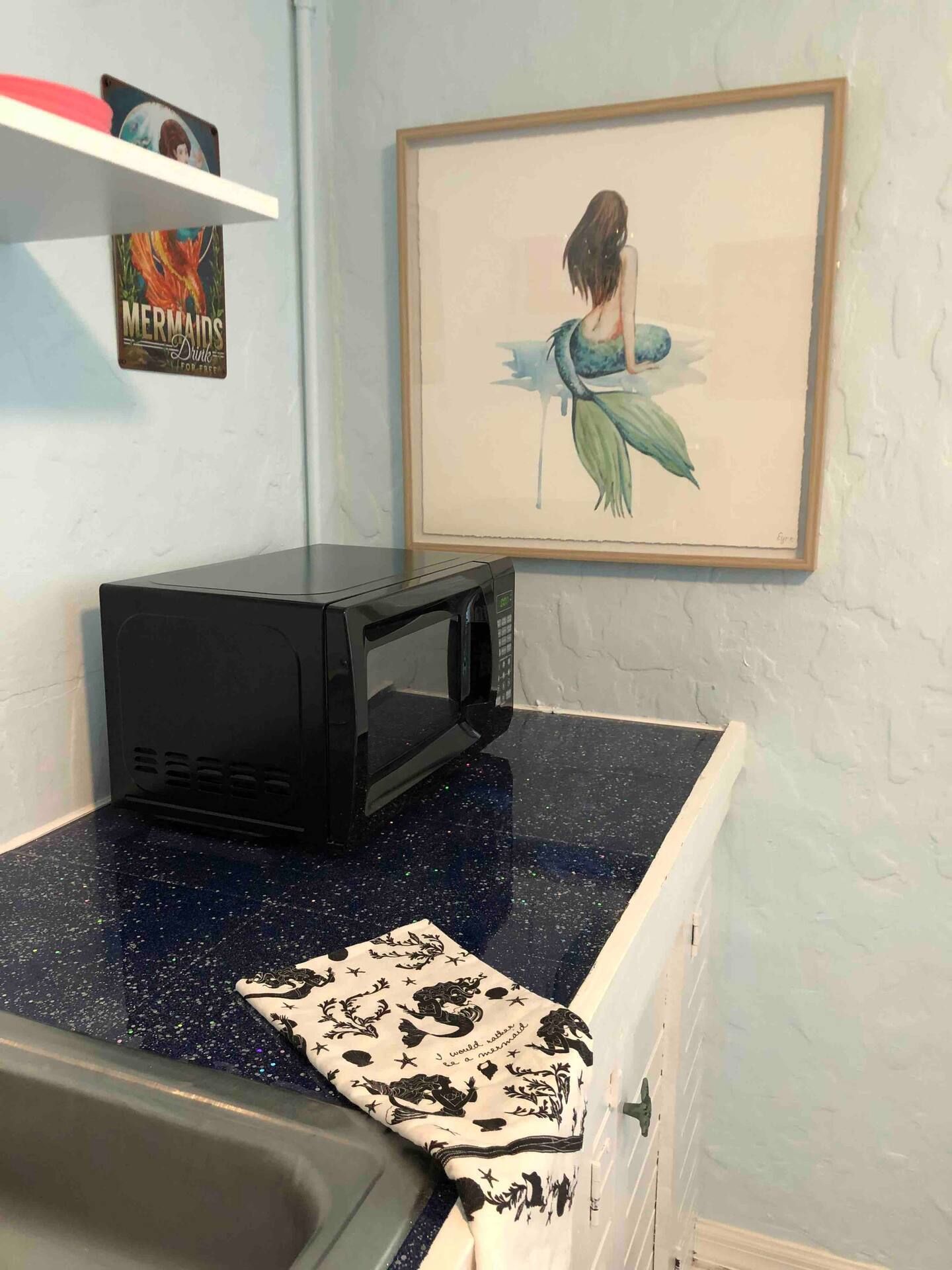 A kitchen counter with a microwave and a picture of a mermaid on the wall.