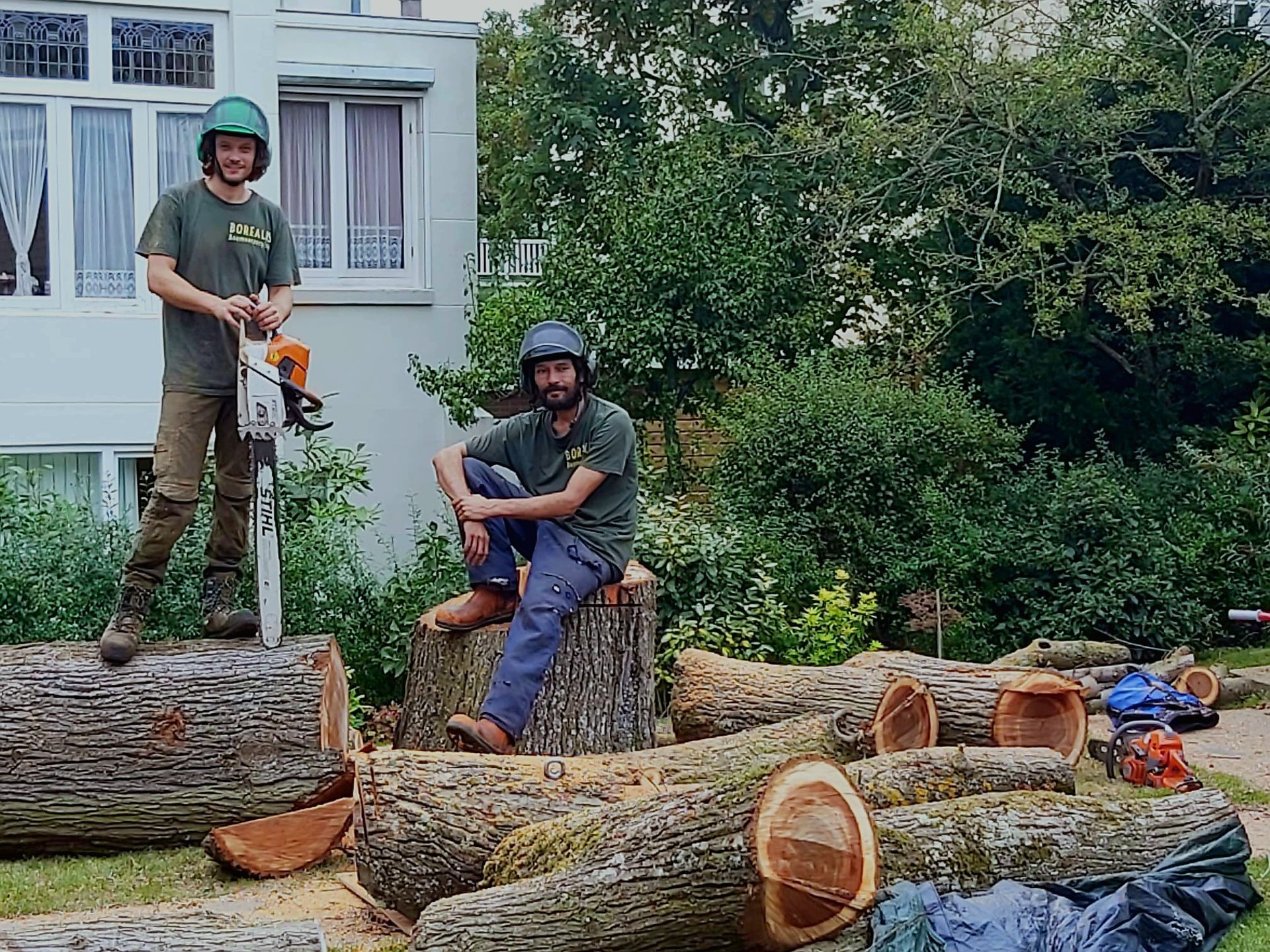 Twee boomverzorgers met helmen poseren buiten bij boomstammen en een kettingzaag.