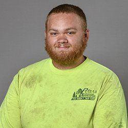 Man with reddish-brown beard in a green shirt, smiling. Shirt has a logo, against a gray background.