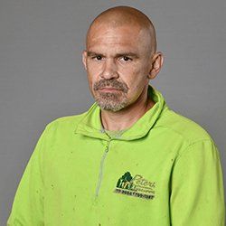 Bald man in green shirt with logo, looking directly at the viewer against a gray backdrop.
