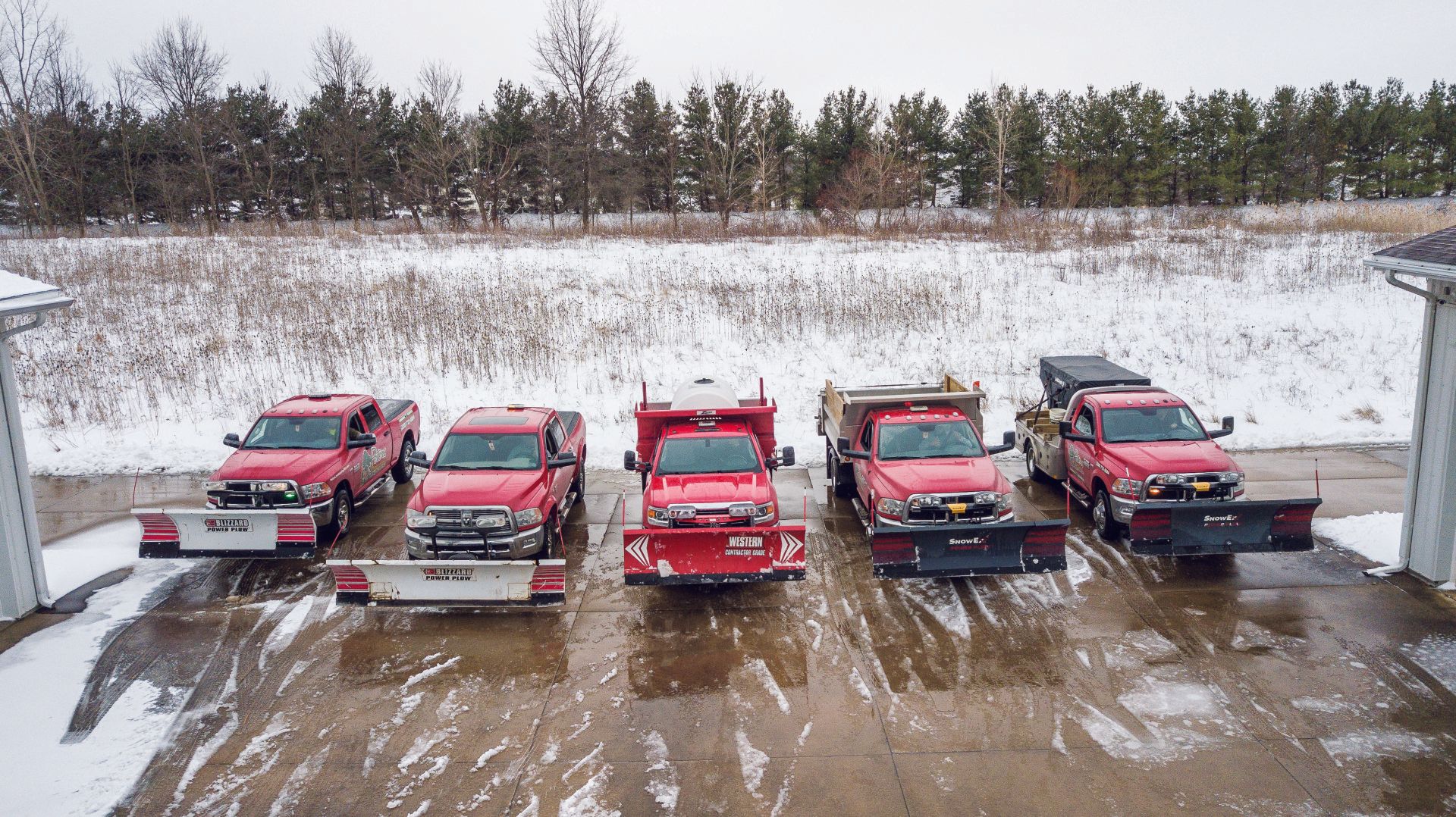 Five red snowplow trucks parked on a snowy driveway, trees in the background.