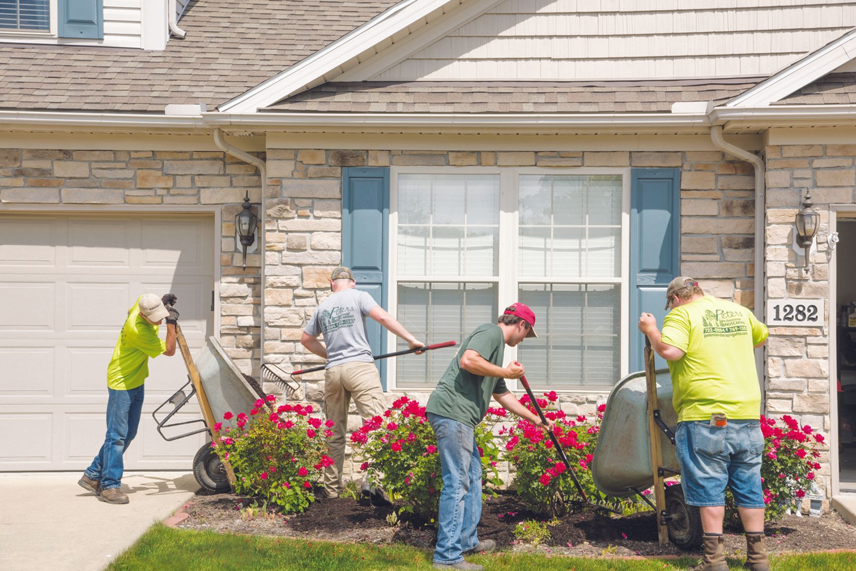 Four landscapers planting flowers in front of a beige house with blue shutters.