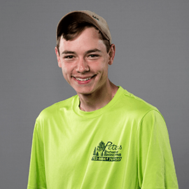 Young man smiling, wearing a tan cap and bright yellow shirt with logo, standing against a gray background.