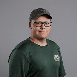 Man in a green shirt and glasses, wearing a cap, smiling at the camera against a gray background.