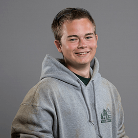 Teenager in a gray hoodie smiles, wearing safety glasses on his head, against a gray background.