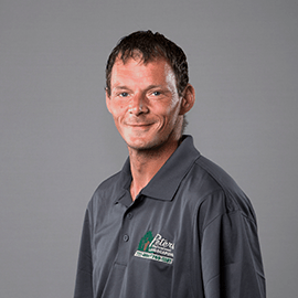 Man wearing gray polo shirt with logo, smiling, against a gray backdrop.