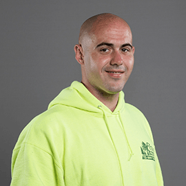 Bald man in a bright yellow hoodie smiles, chest-level portrait against a gray backdrop.