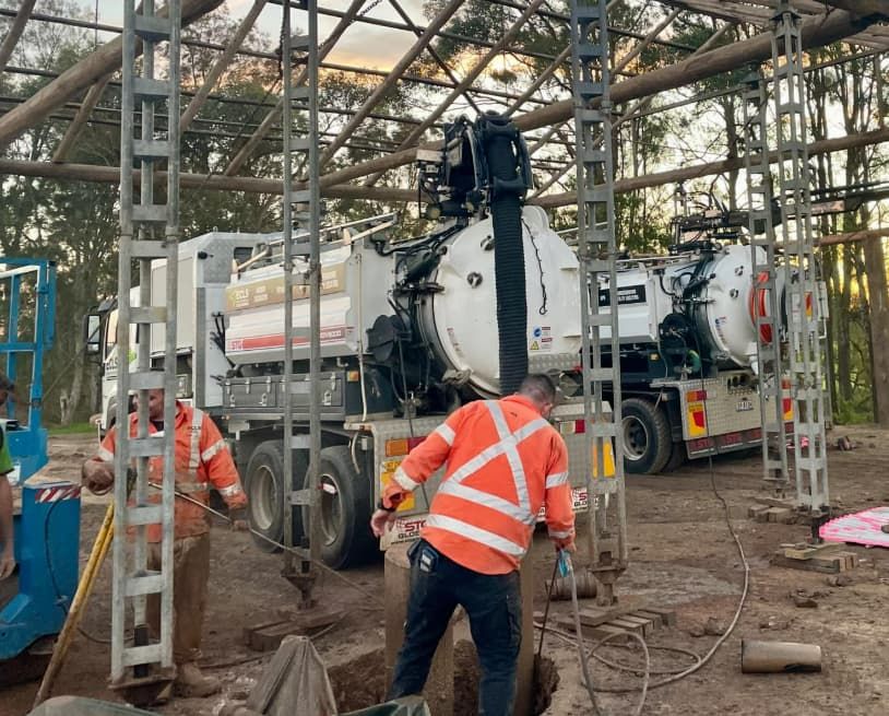 A Man Is Digging A Hole In The Ground In Front Of A Vacuum Truck — East Coast Locating Services In Wallalong, NSW