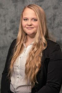 A woman with long blonde hair is wearing a black jacket and a white shirt.