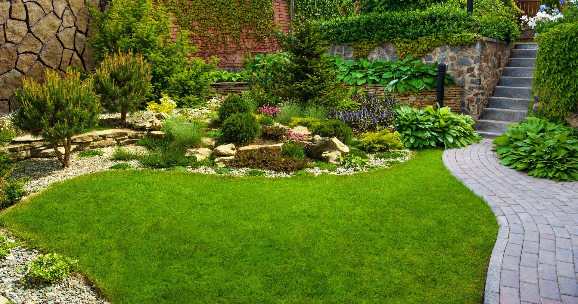 A lush green garden with a brick walkway leading to a stone wall and stairs.