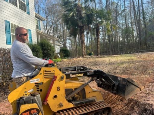 A man is driving a yellow tractor in a yard in front of a house.