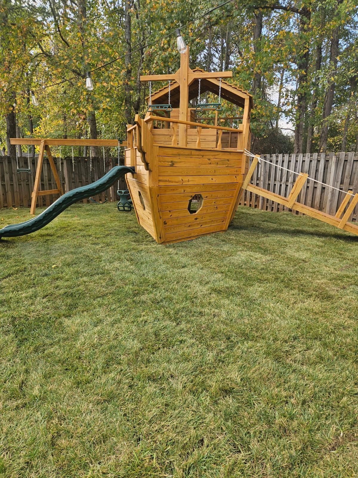A wooden pirate ship playground with a slide and swings in a backyard.