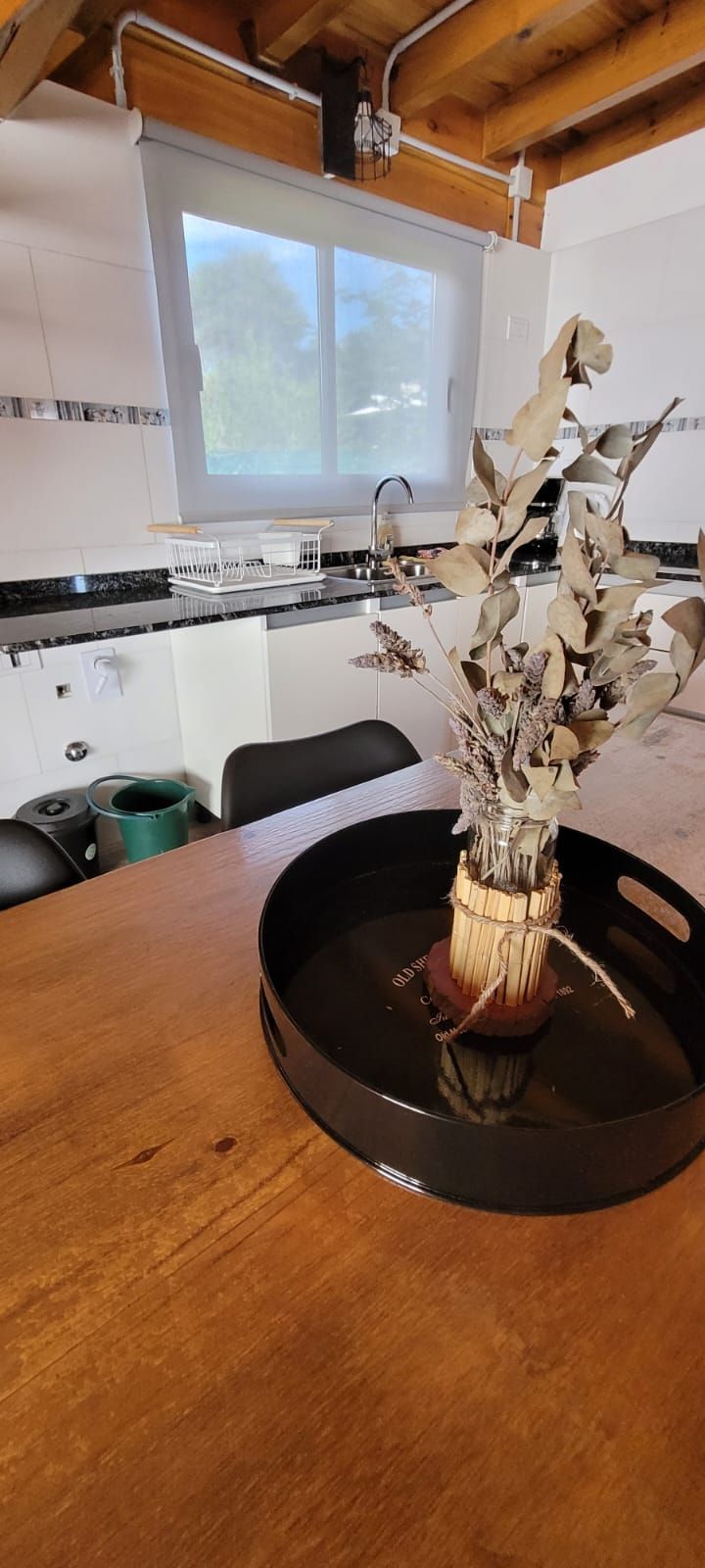 Una mesa de madera con una bandeja negra que contiene un arreglo de plantas secas. Al fondo, una ventana y una encimera de cocina. Cabaña Los Ciruelos