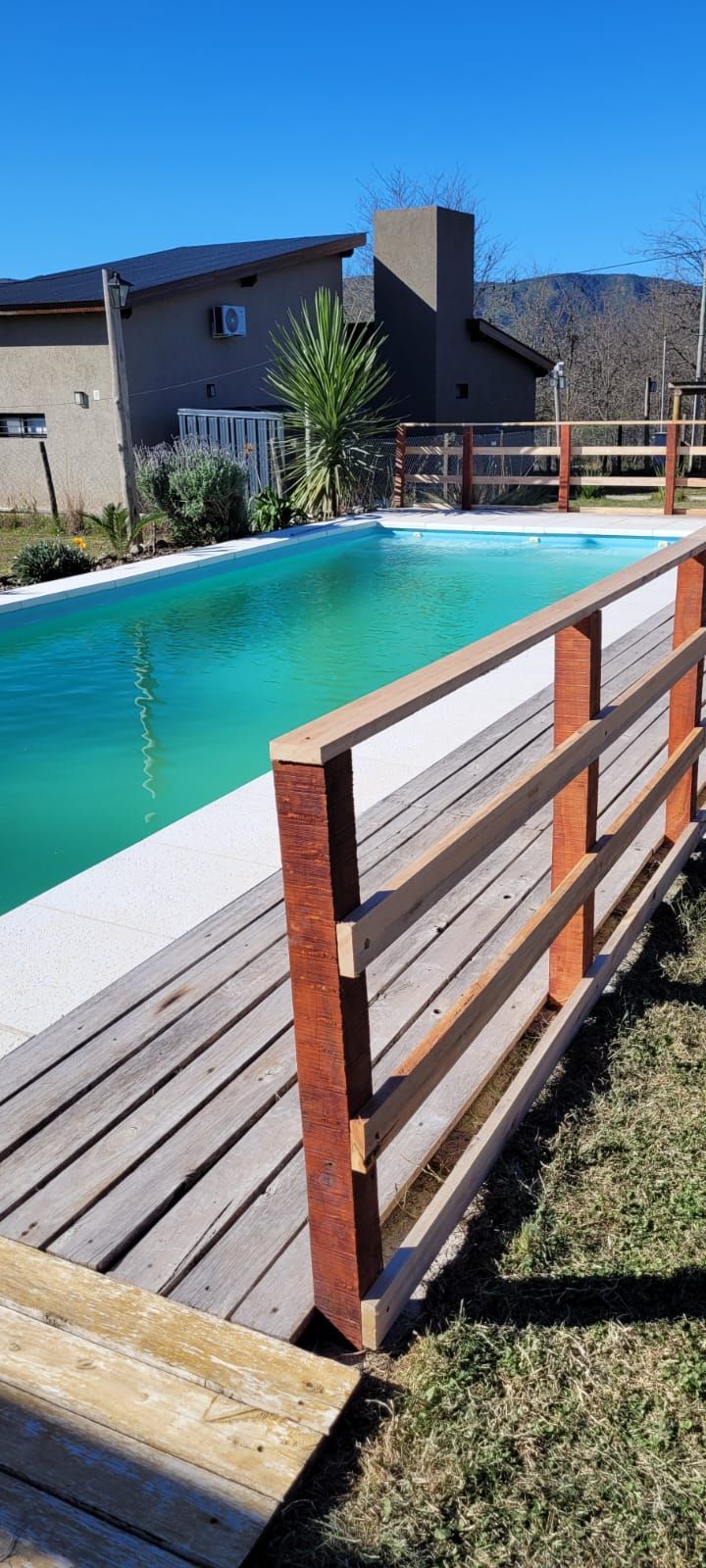 Una piscina de agua turquesa junto a una terraza y una valla de madera. Al fondo se ve una cabaña. Cabaña Los Ciruelos