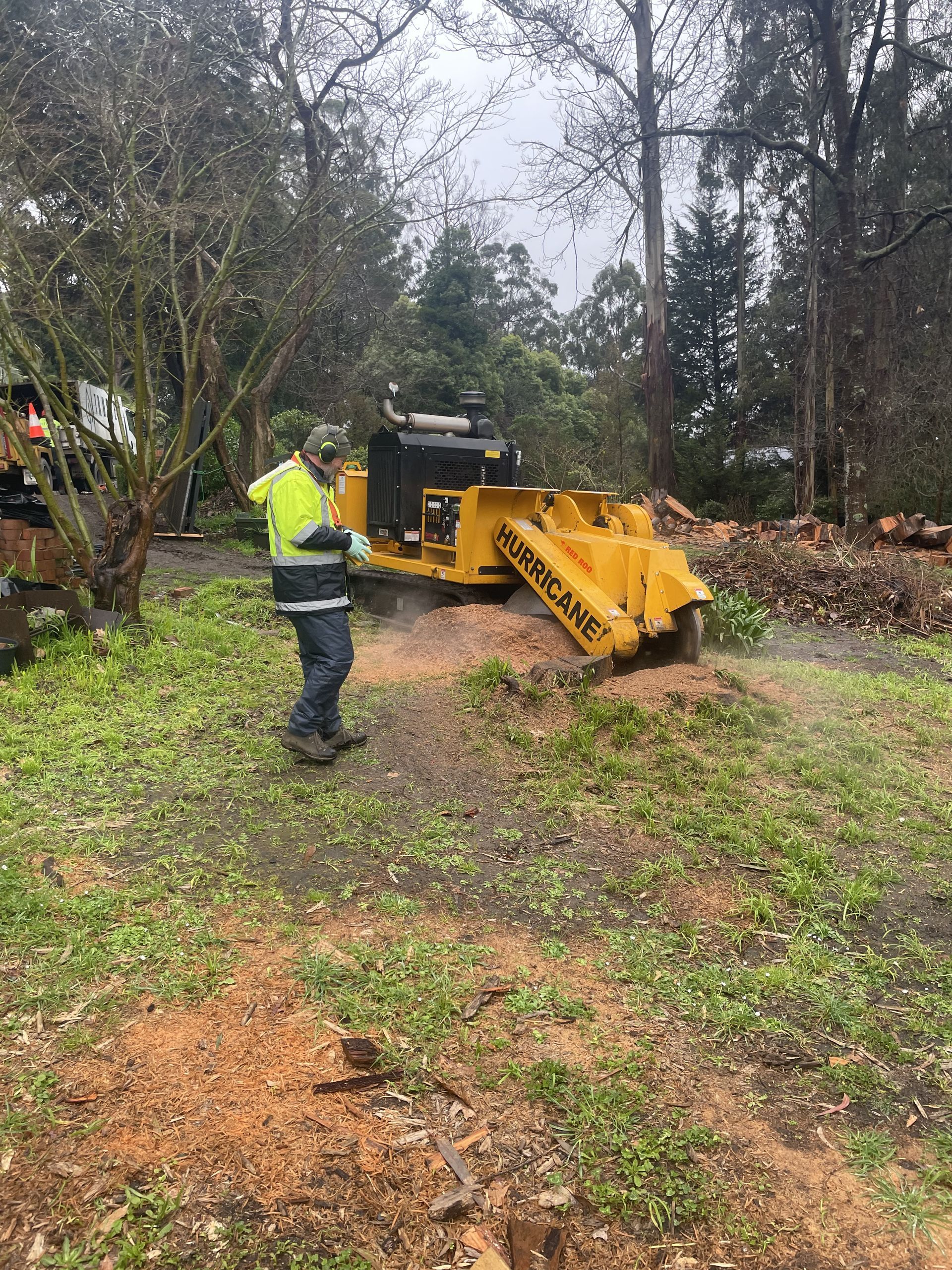 Worker in a neon jacket using a yellow wood chipper on a muddy wooded path.