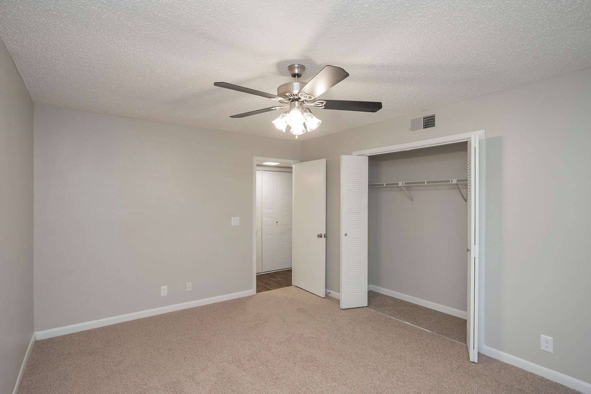 An empty, neutral-colored bedroom featuring a ceiling fan, beige carpet, and open closet doors.
