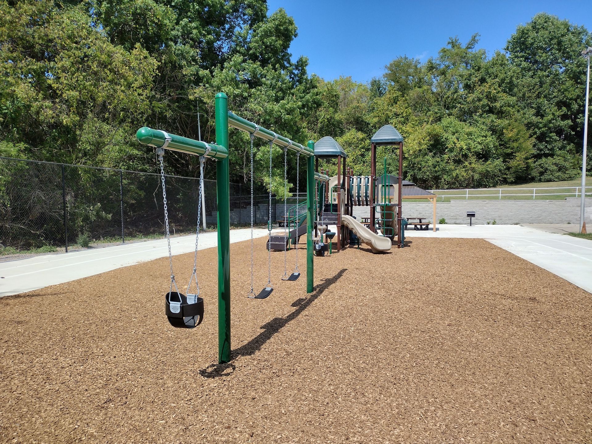 A playground with a swing set in the foreground and a climbing structure with a slide on a wood-chip ground covering.