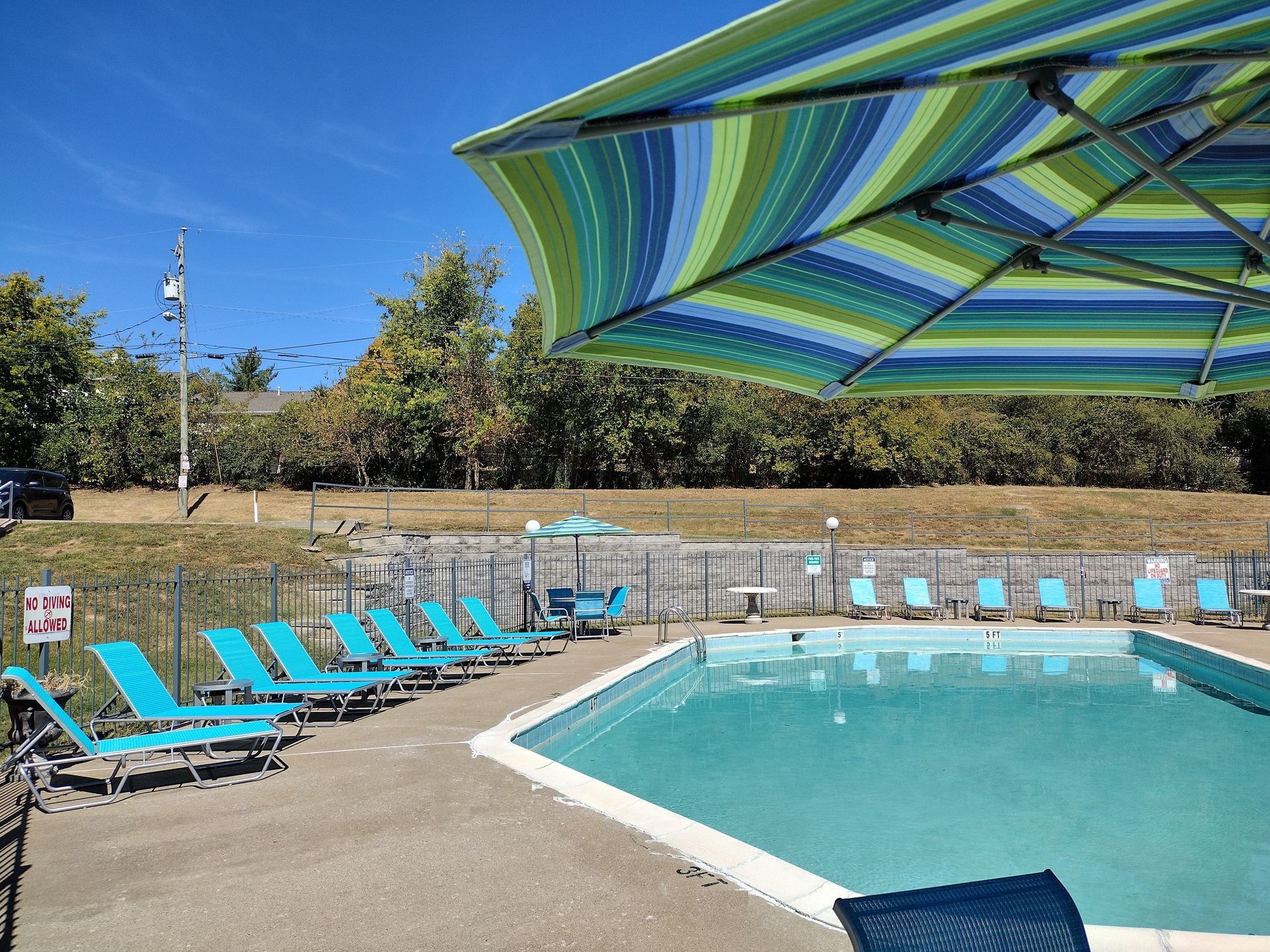 A swimming pool surrounded by teal lounge chairs under a large, striped blue and green umbrella on a sunny day.