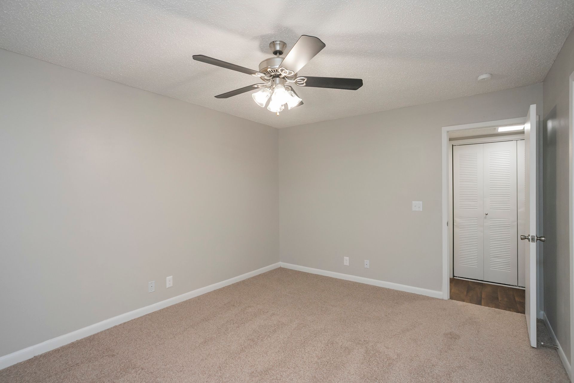 An empty bedroom with light gray walls, tan carpet, a ceiling fan, and an open door revealing a white louvered closet.