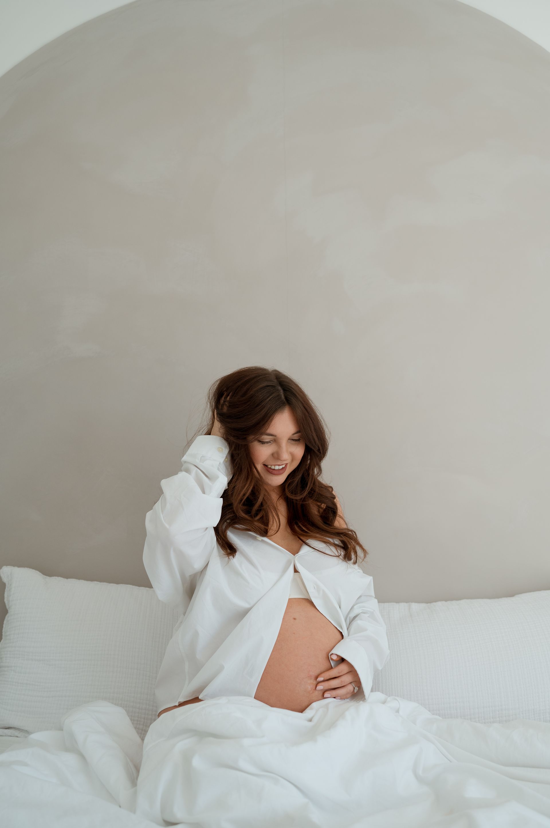A pregnant woman is sitting on a bed with white sheets.