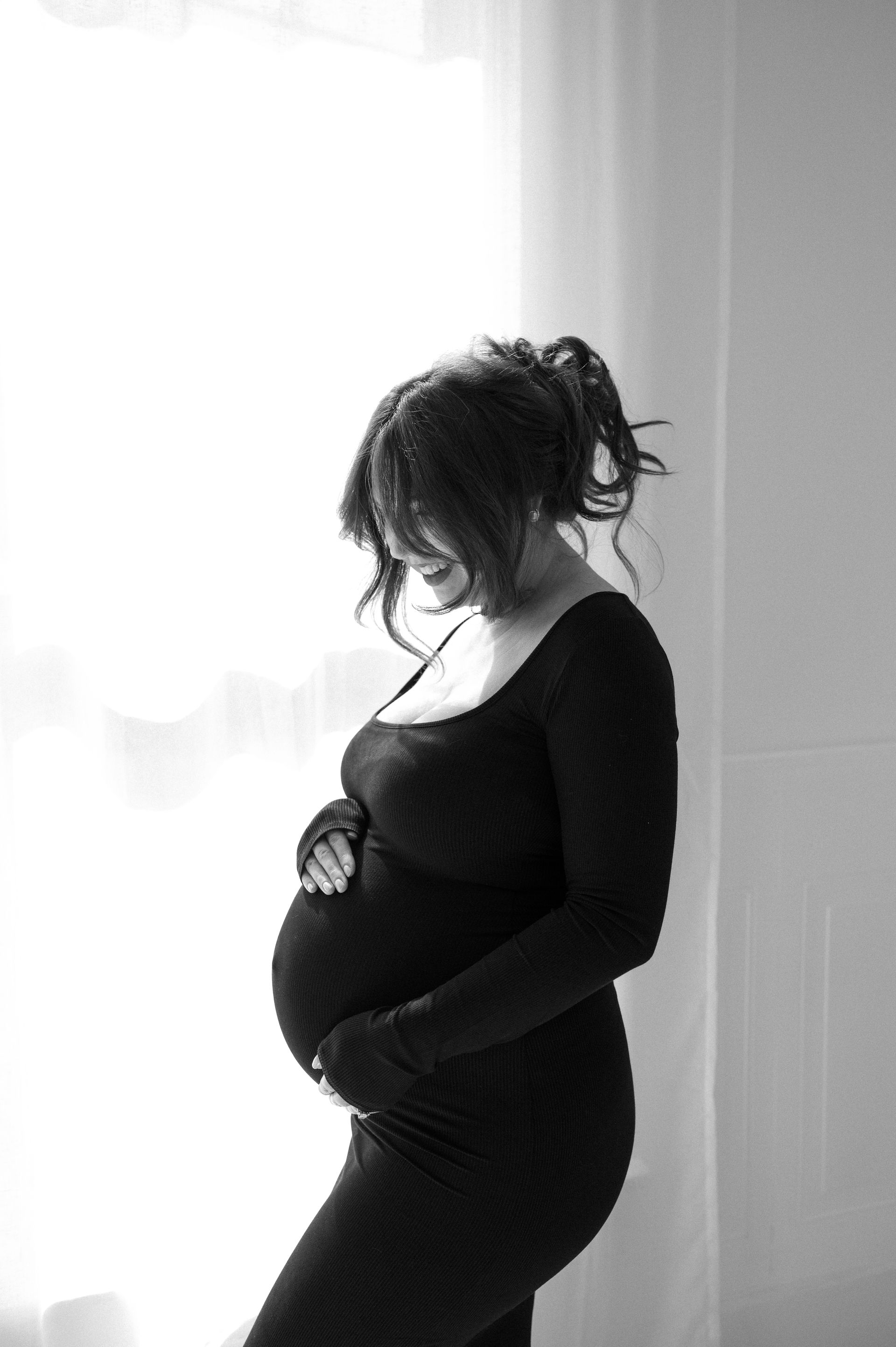 A pregnant woman in a black dress is standing in front of a window holding her belly.