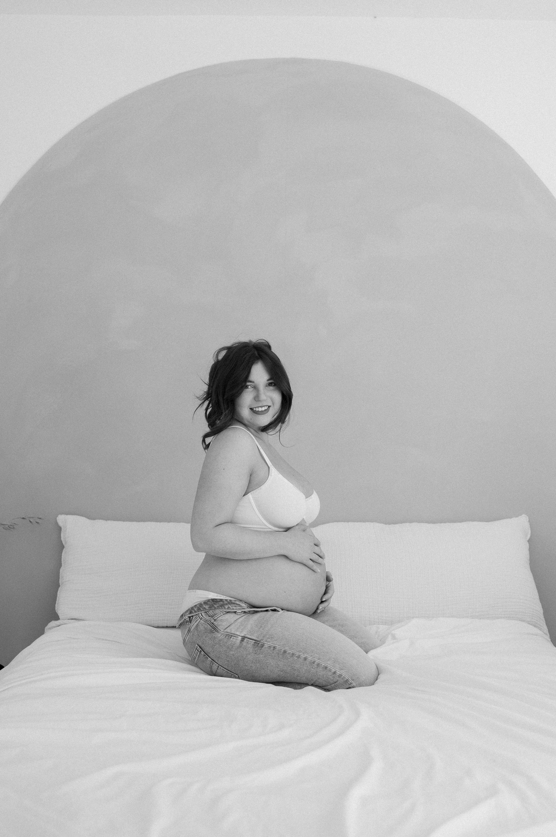 A pregnant woman is sitting on a bed in a black and white photo.