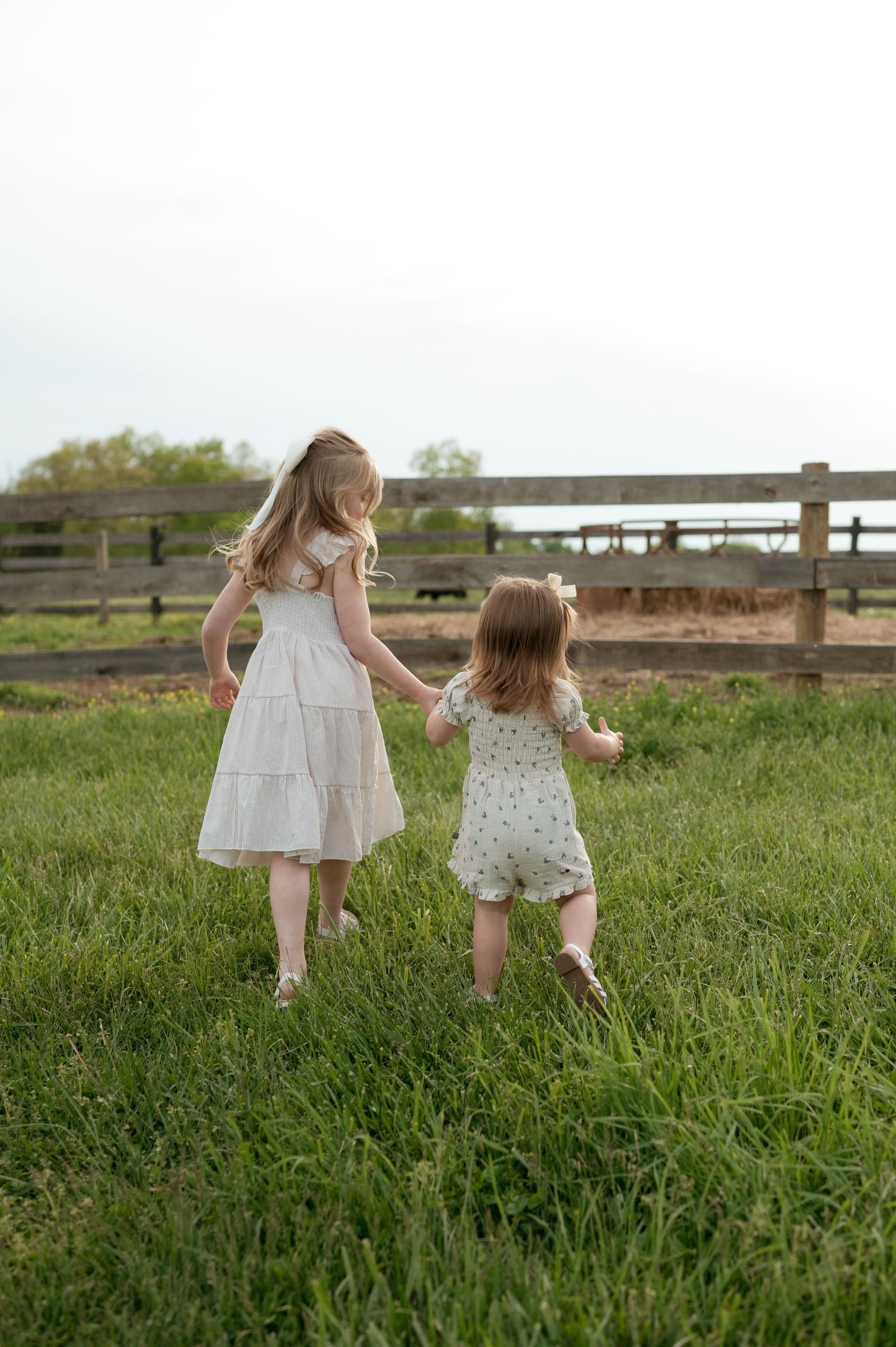 Two little girls are holding hands and running through a grassy field.