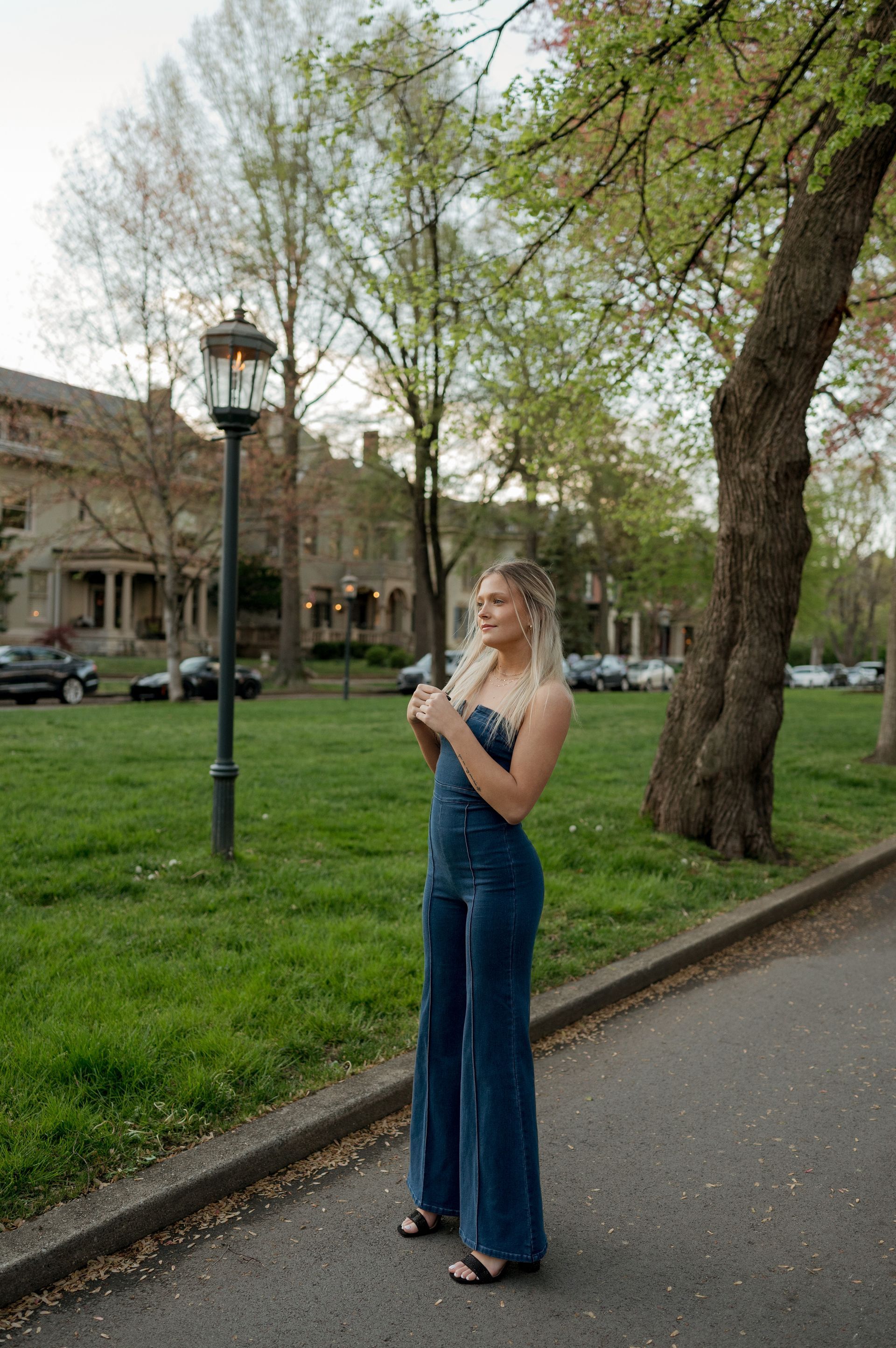 A woman in a blue jumpsuit is standing on a sidewalk in a park.