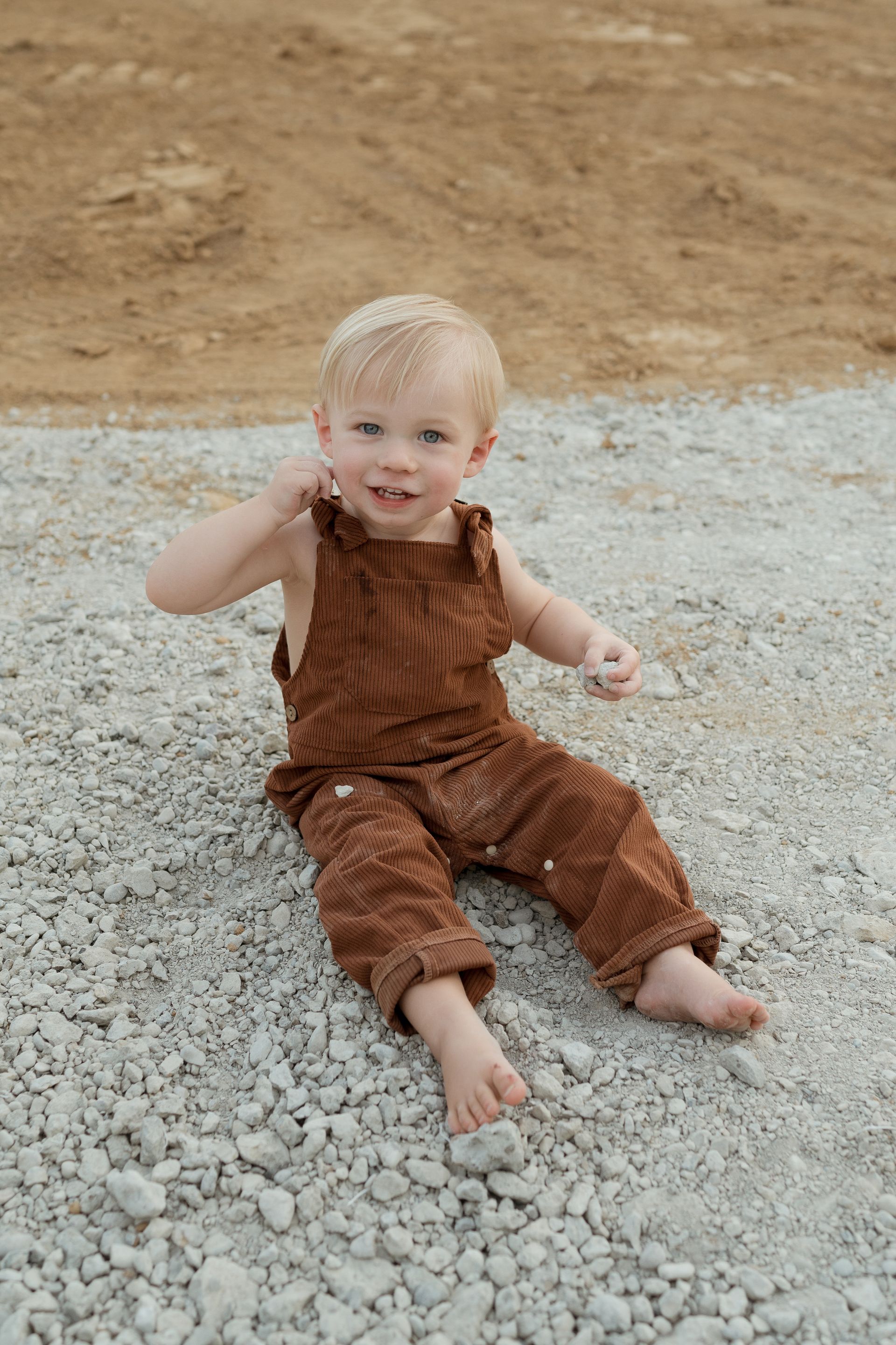 A little boy in brown overalls is sitting on a pile of gravel.