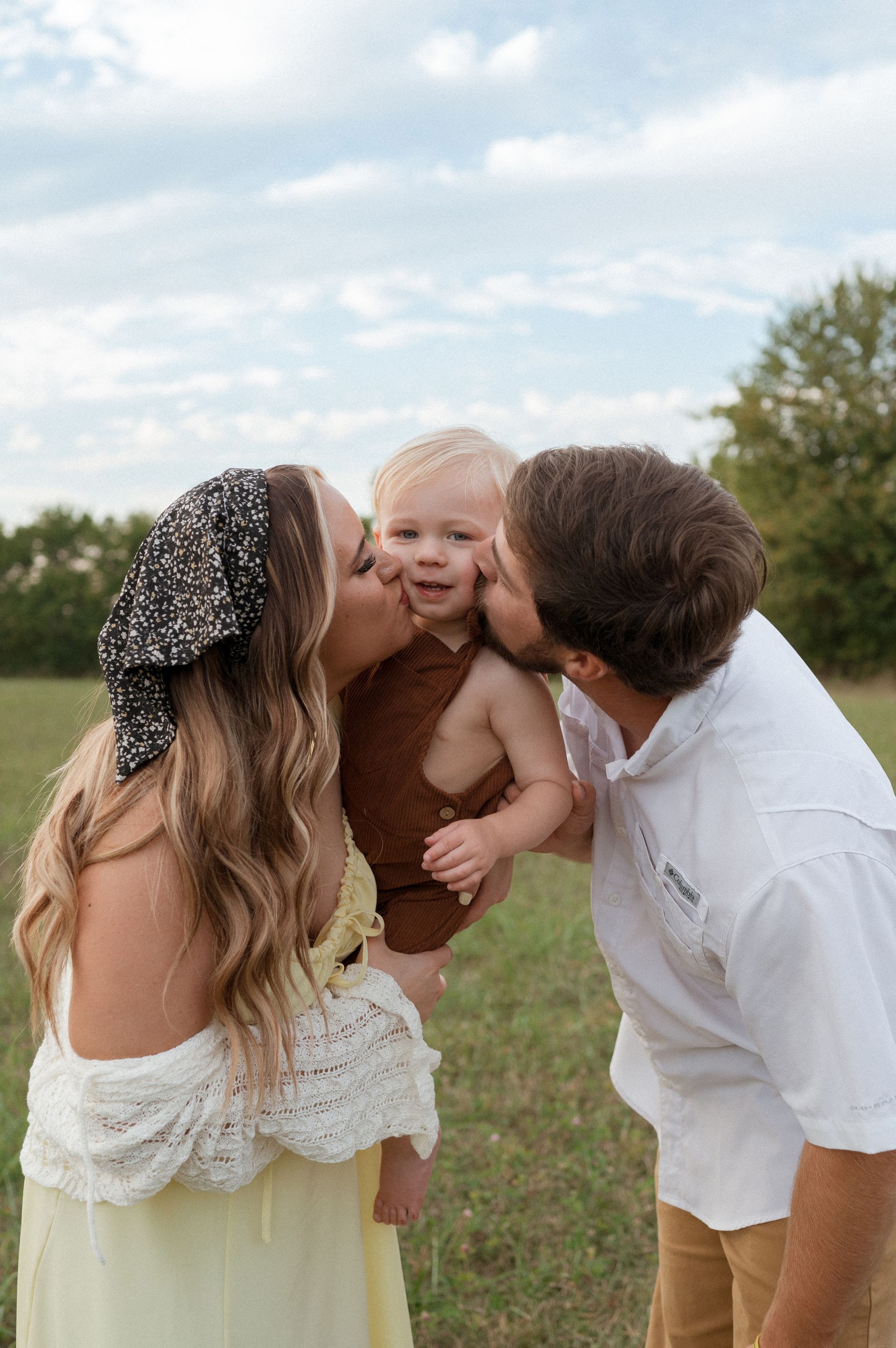 A man and a woman are kissing a baby on the cheek in a field.