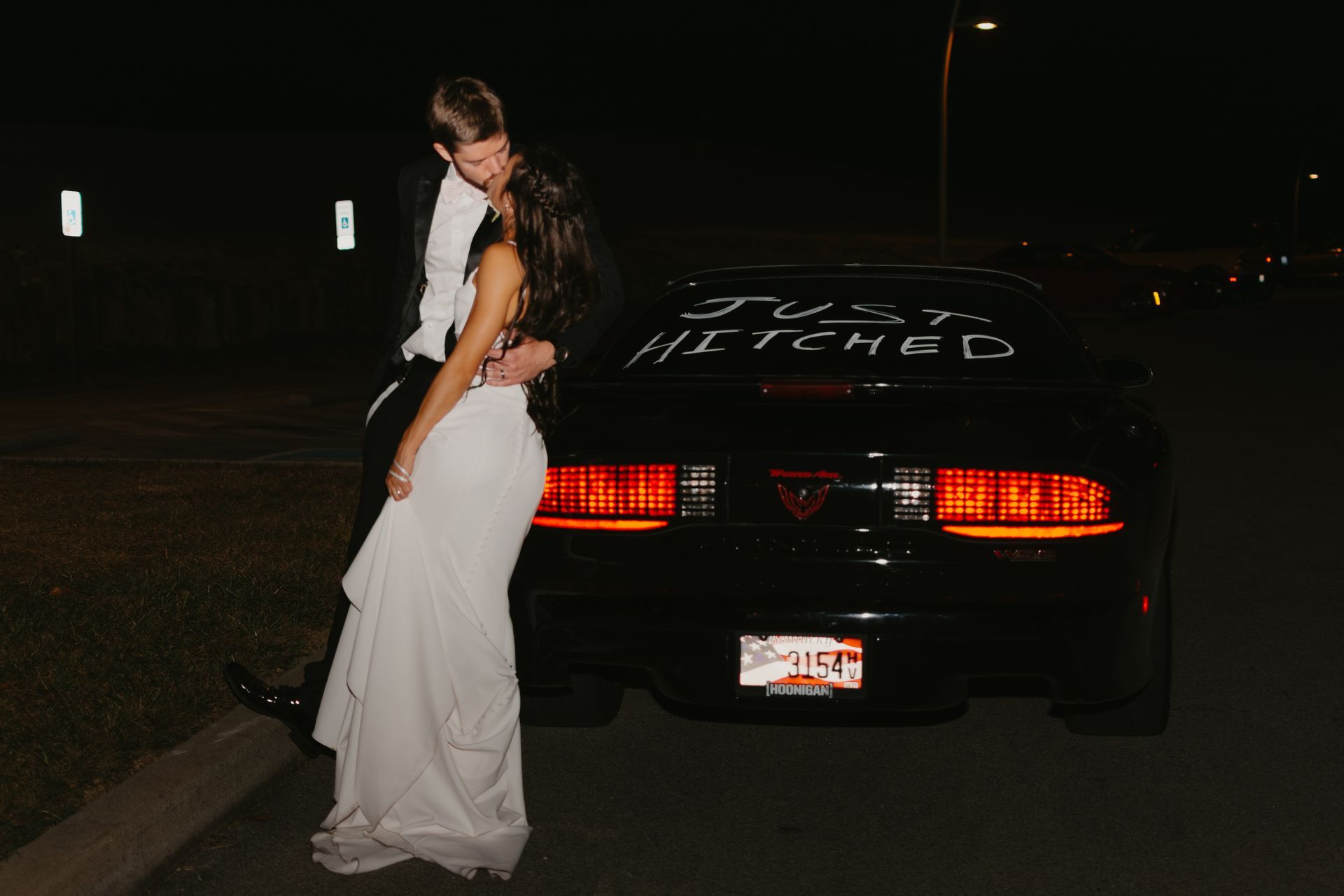 A bride and groom are kissing in front of a black car.