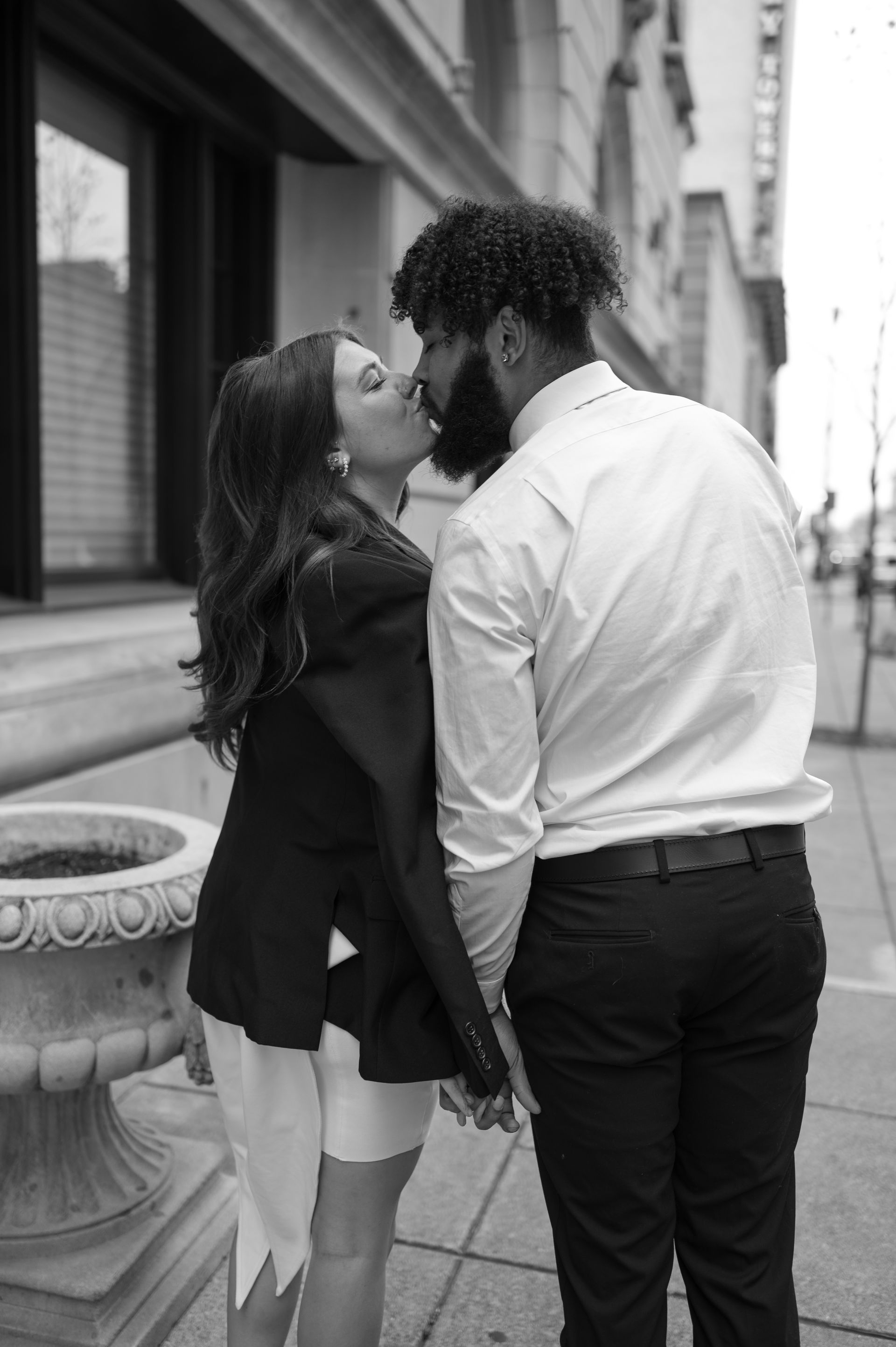 A black and white photo of a man and woman kissing on a sidewalk.