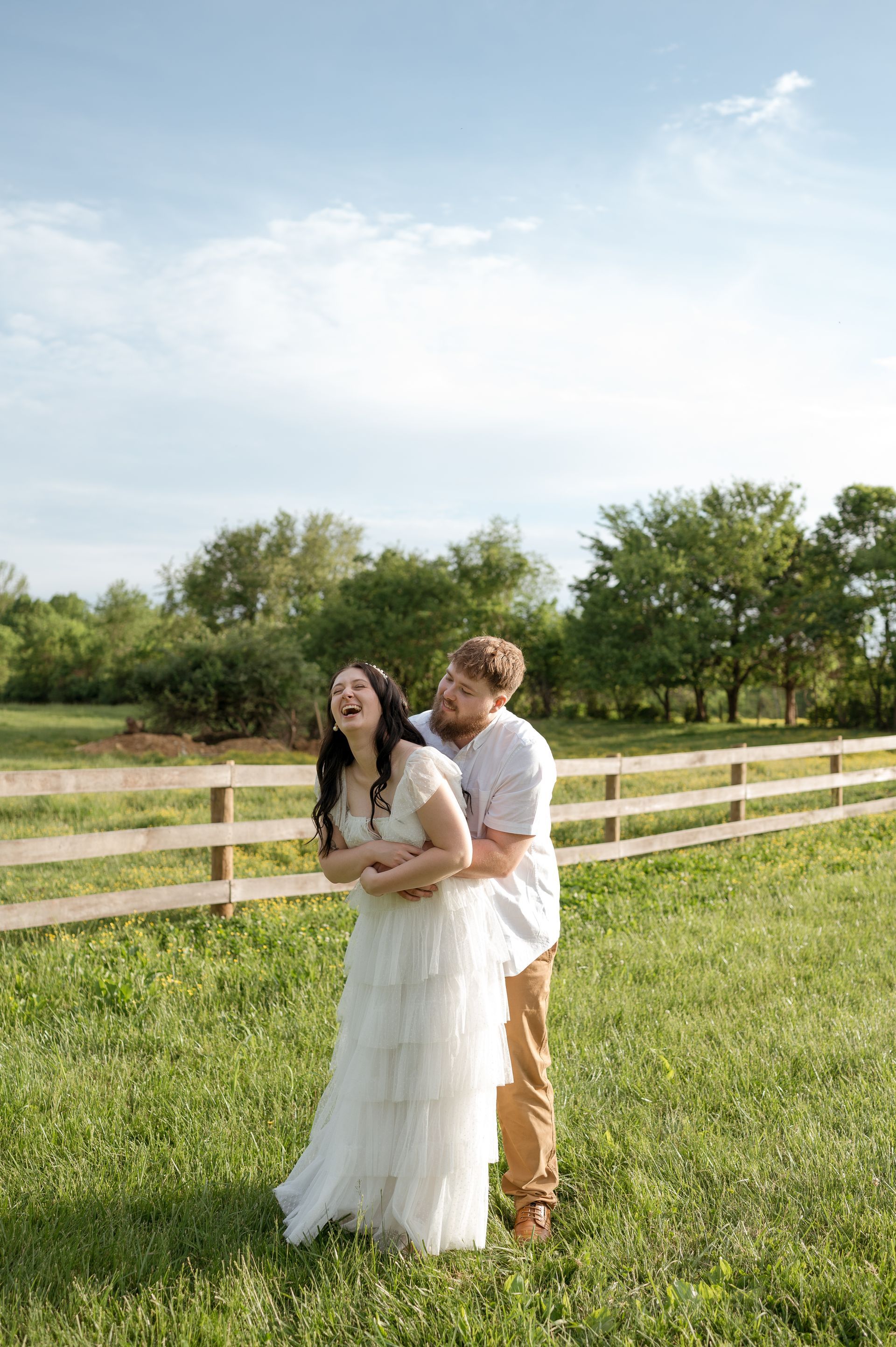 A bride and groom are laughing in a field next to a wooden fence.