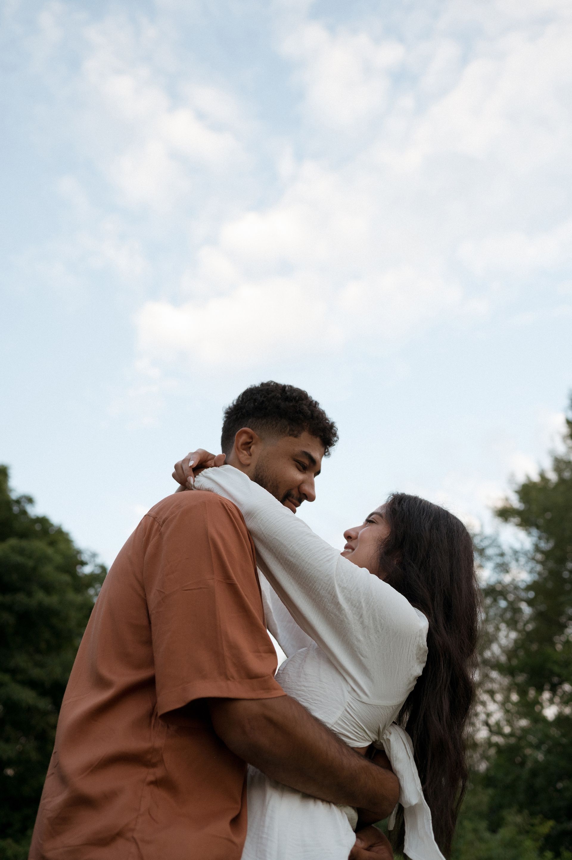 A man and a woman are hugging each other in front of a cloudy sky.