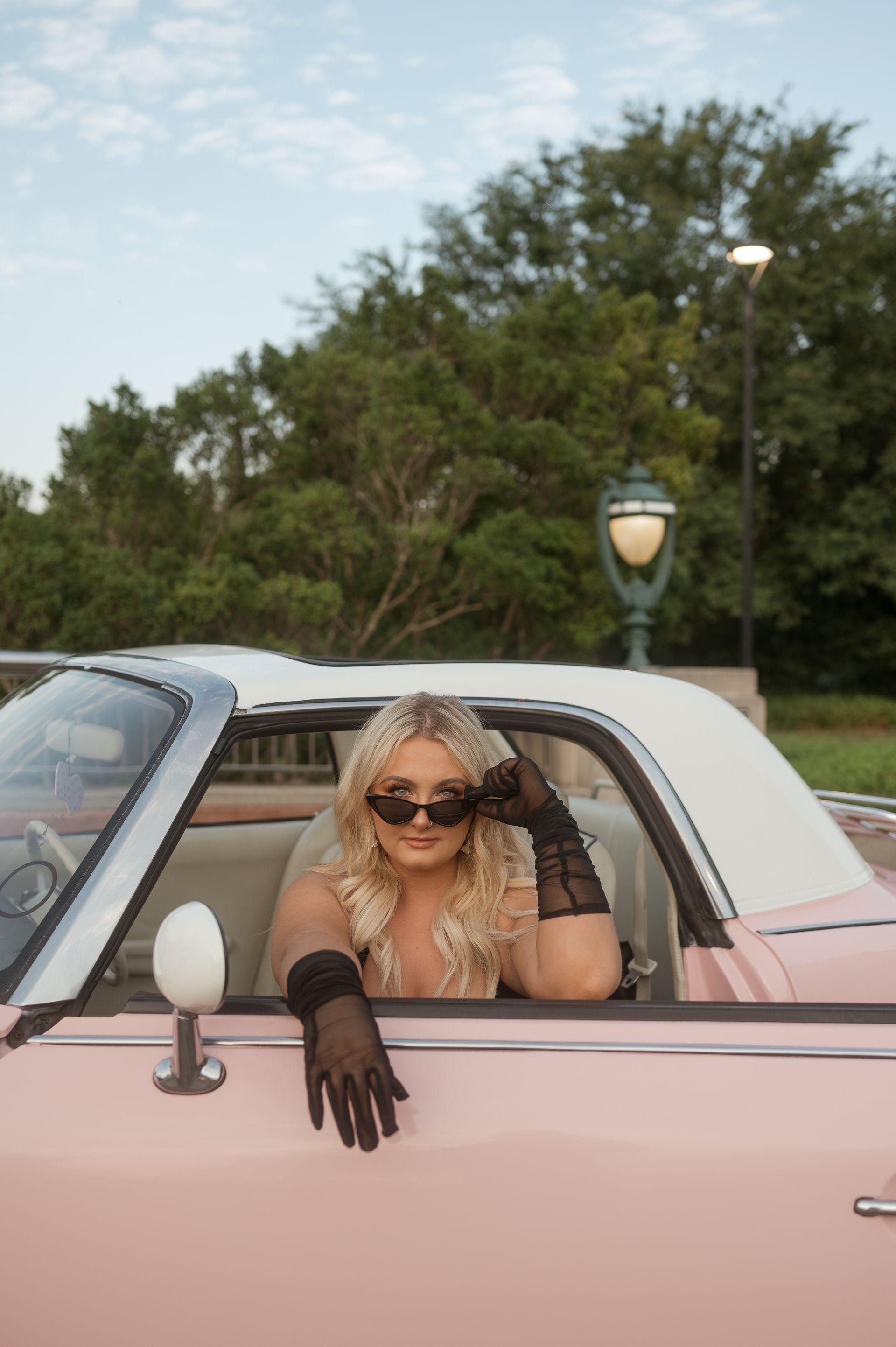 A woman wearing sunglasses and gloves is sitting in a pink car.