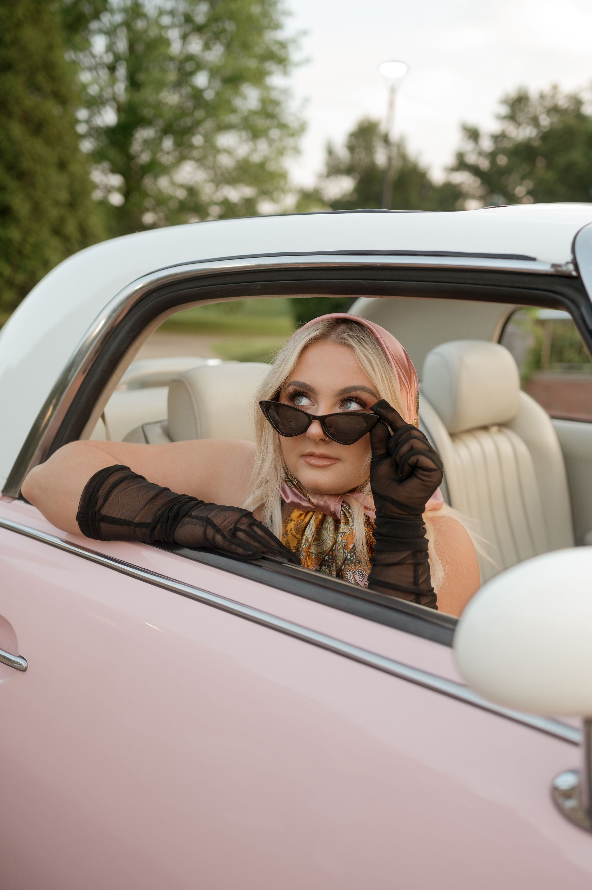 A woman wearing sunglasses and gloves is sitting in a pink car.