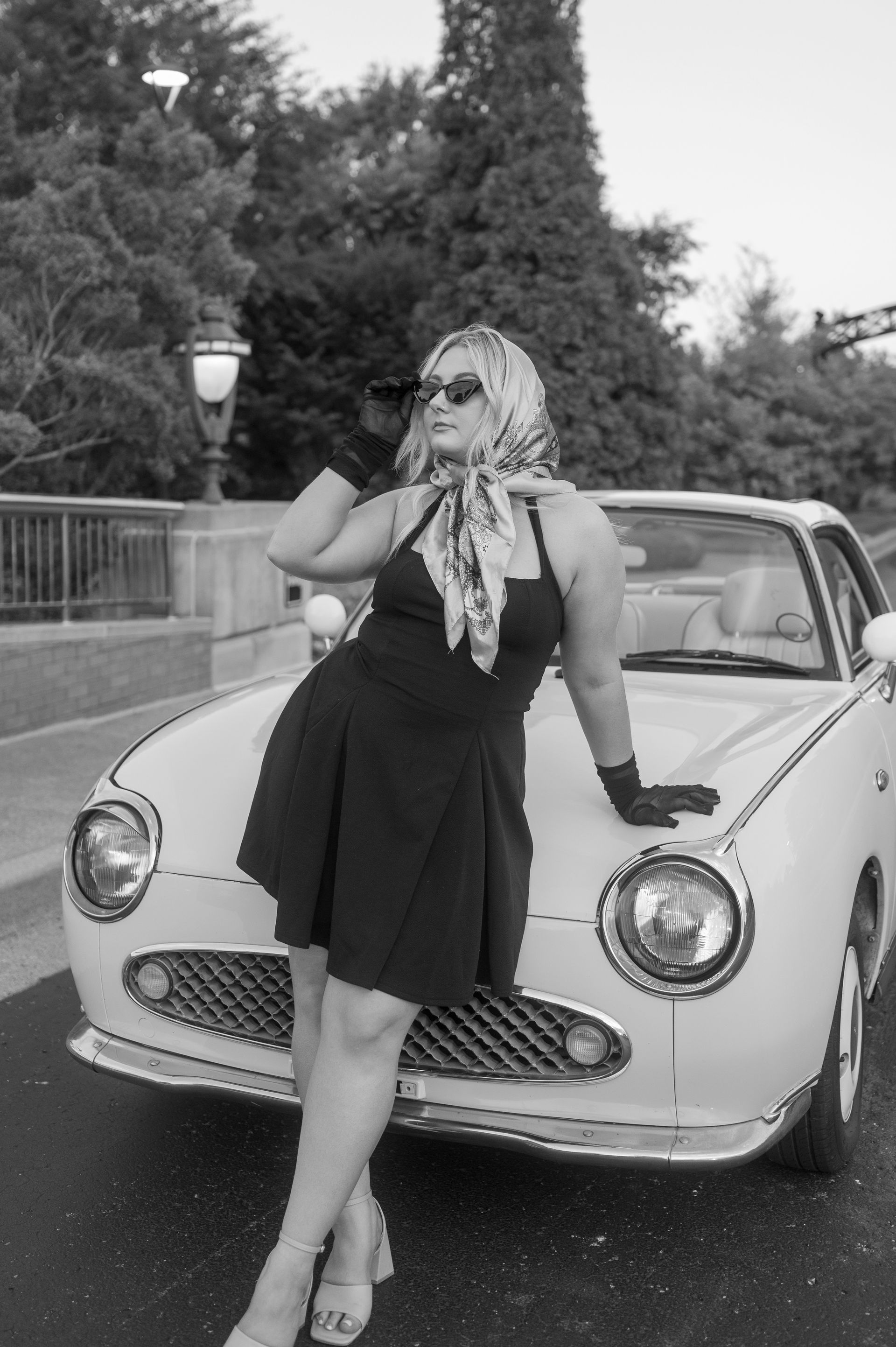 A woman in a black dress leaning against a white car